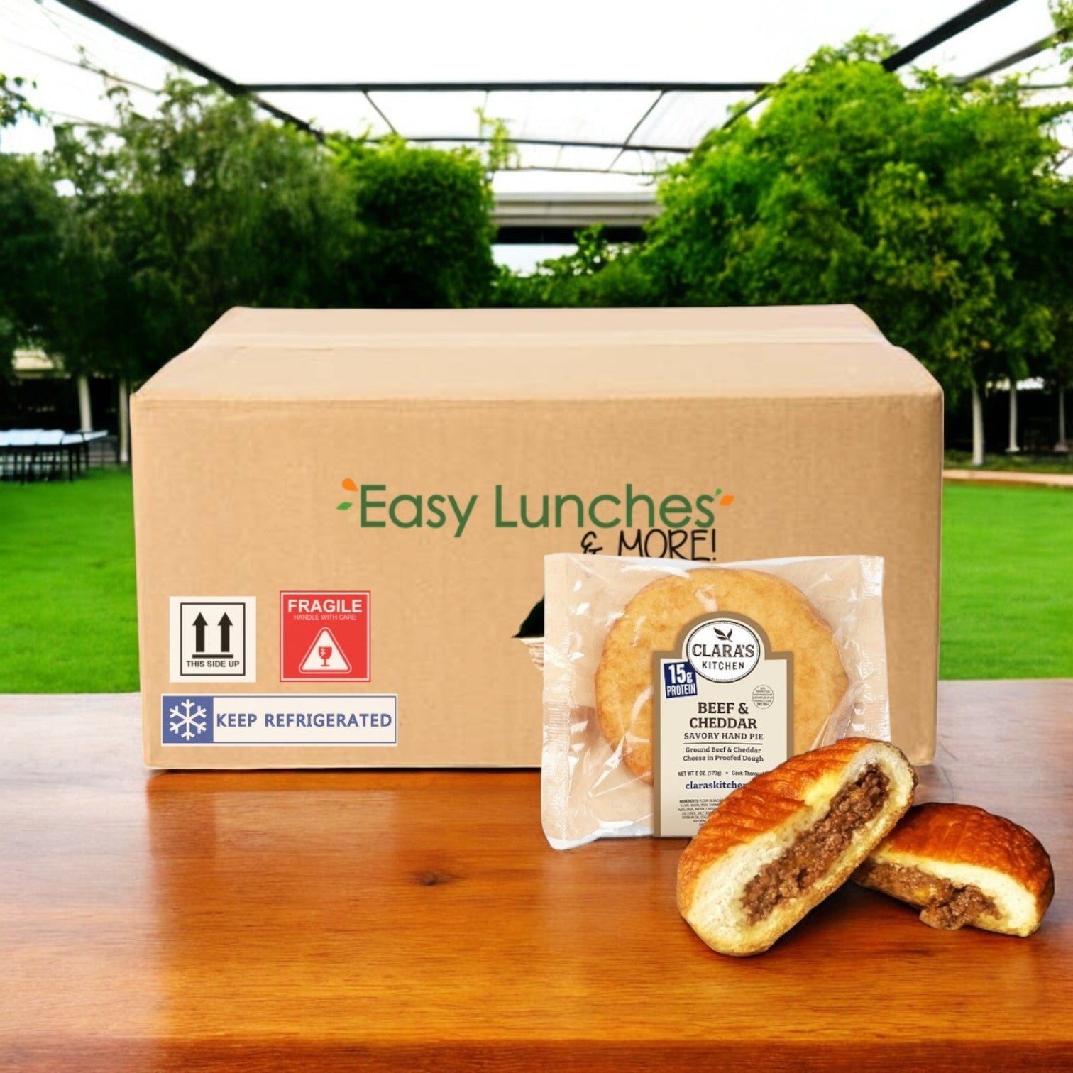 A box labeled "Easy Lunches & More" sits outdoors featuring Clara's Kitchen Beef & Cheddar Handpie, with its flaky pastry. In front is a sealed handpie and a halved version showcasing flavorful seasoned ground beef.