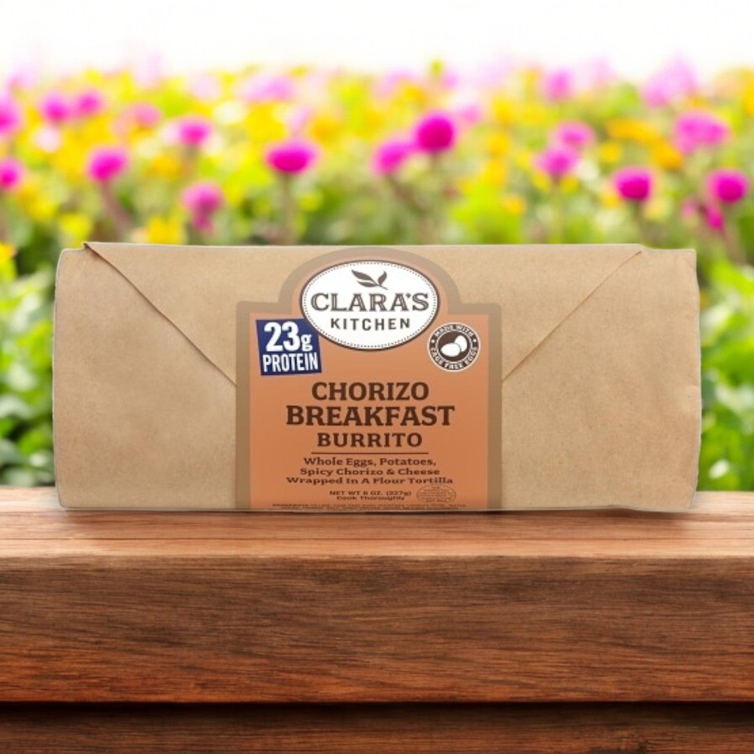 A Clara's Kitchen 8 oz Individually Packaged Sausage Breakfast Burrito, with 23g of protein, rests on a wooden surface. Blurred colorful flowers in the background add a whimsical touch to this grab-and-go meal.