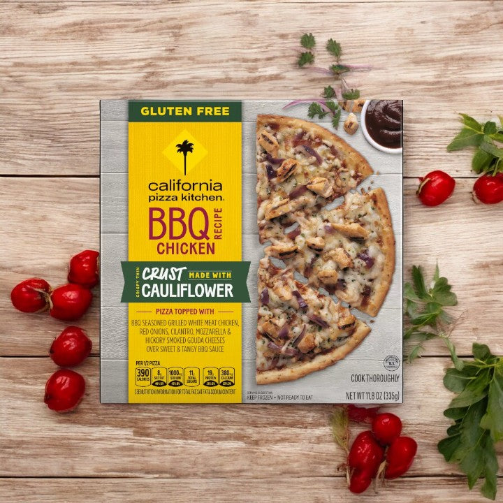 A packaged California Pizza Kitchen Cauliflower Crust BBQ Recipe Chicken pizza, 11.8 oz. - 1 Count, from California Pizza Kitchen is placed on a wooden surface surrounded by fresh cherry tomatoes and herbs.
