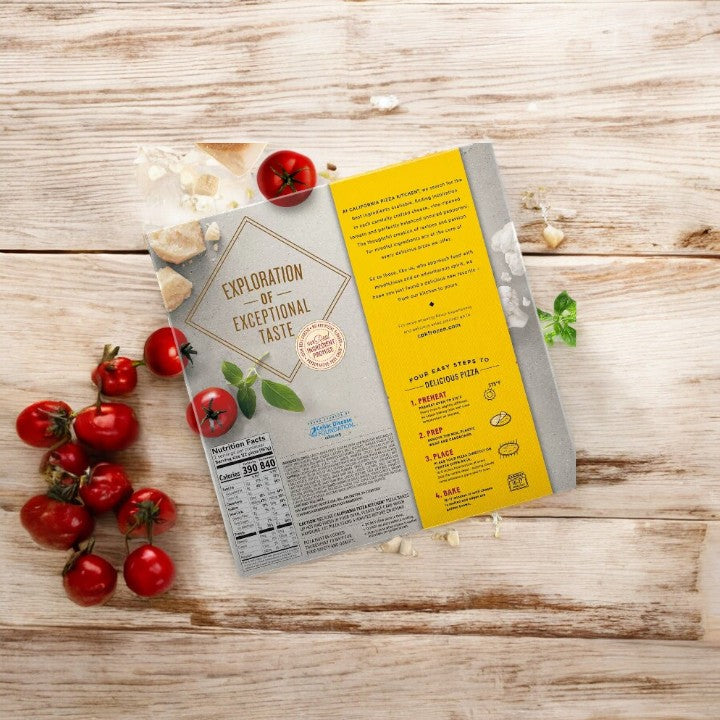 Image of an open pizza box featuring a printed message on a wooden table with scattered ingredients like cherry tomatoes, garlic, and basil. The box's text reads "Exploration of Exceptional Taste," highlighting the deliciousness of our California Pizza Kitchen Cauliflower Crust BBQ Recipe Chicken pizza.