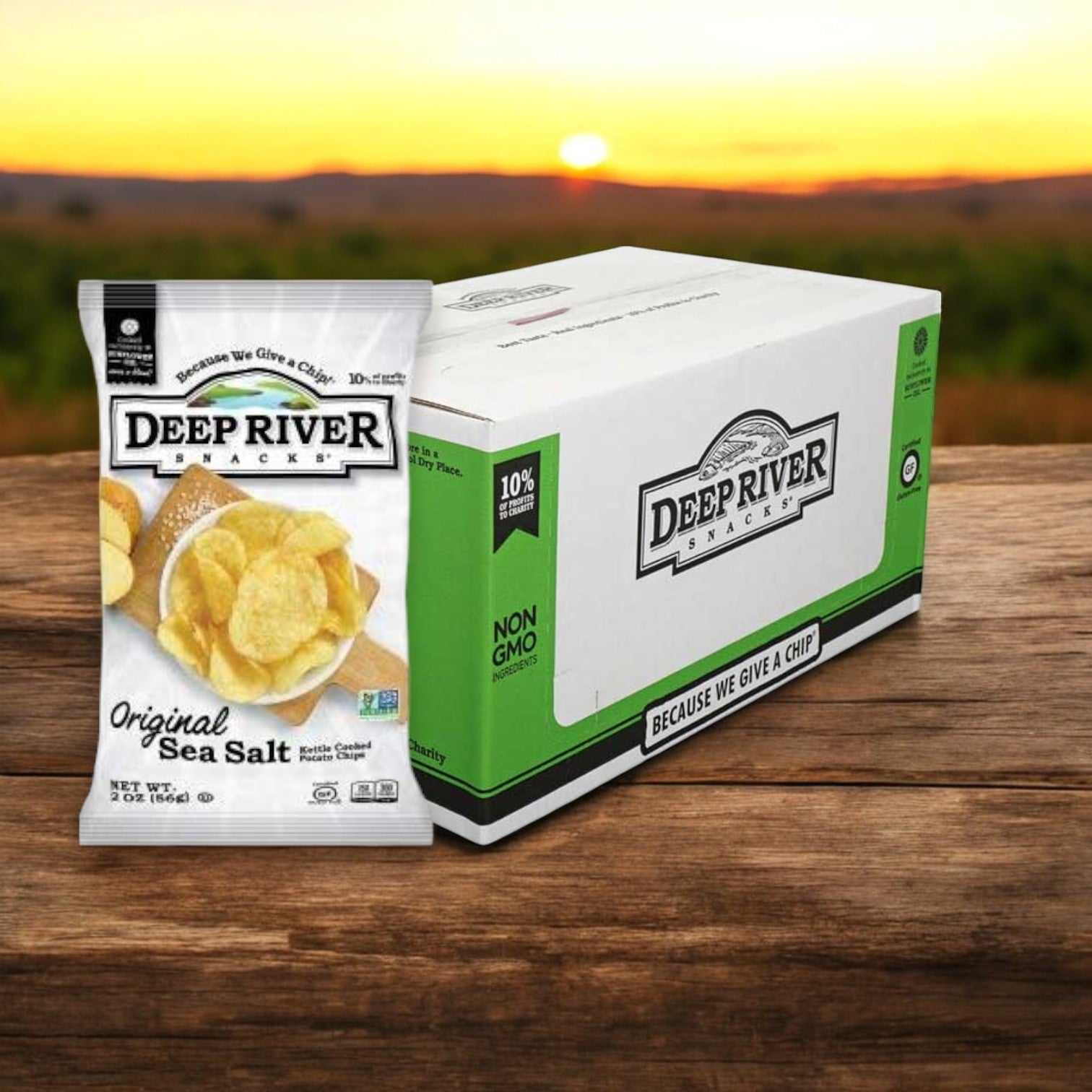 A bag and box of Deep River Snacks - Original Sea Salt Kettle Potato Chips (2 oz, 24 per case) sit on a wooden surface, against a sunset and field backdrop, highlighting these tasty gluten-free snacks.
