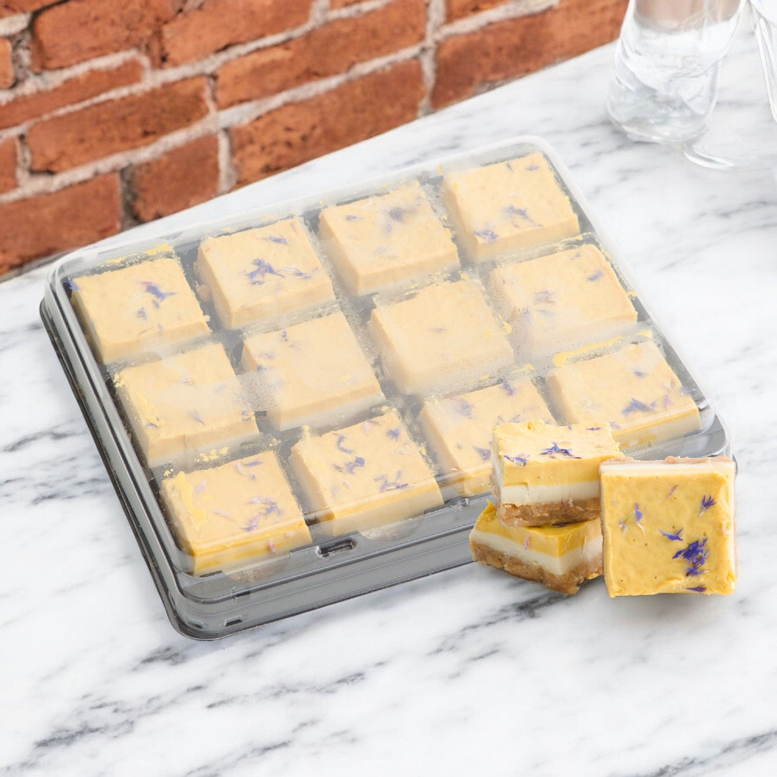 A tray of Nats Vegan Cakes, 12 square yellow-frosted lime mango desserts, sits on a marble surface against a brick wall, with two pieces stacked on the side for a delightful plant-based treat.