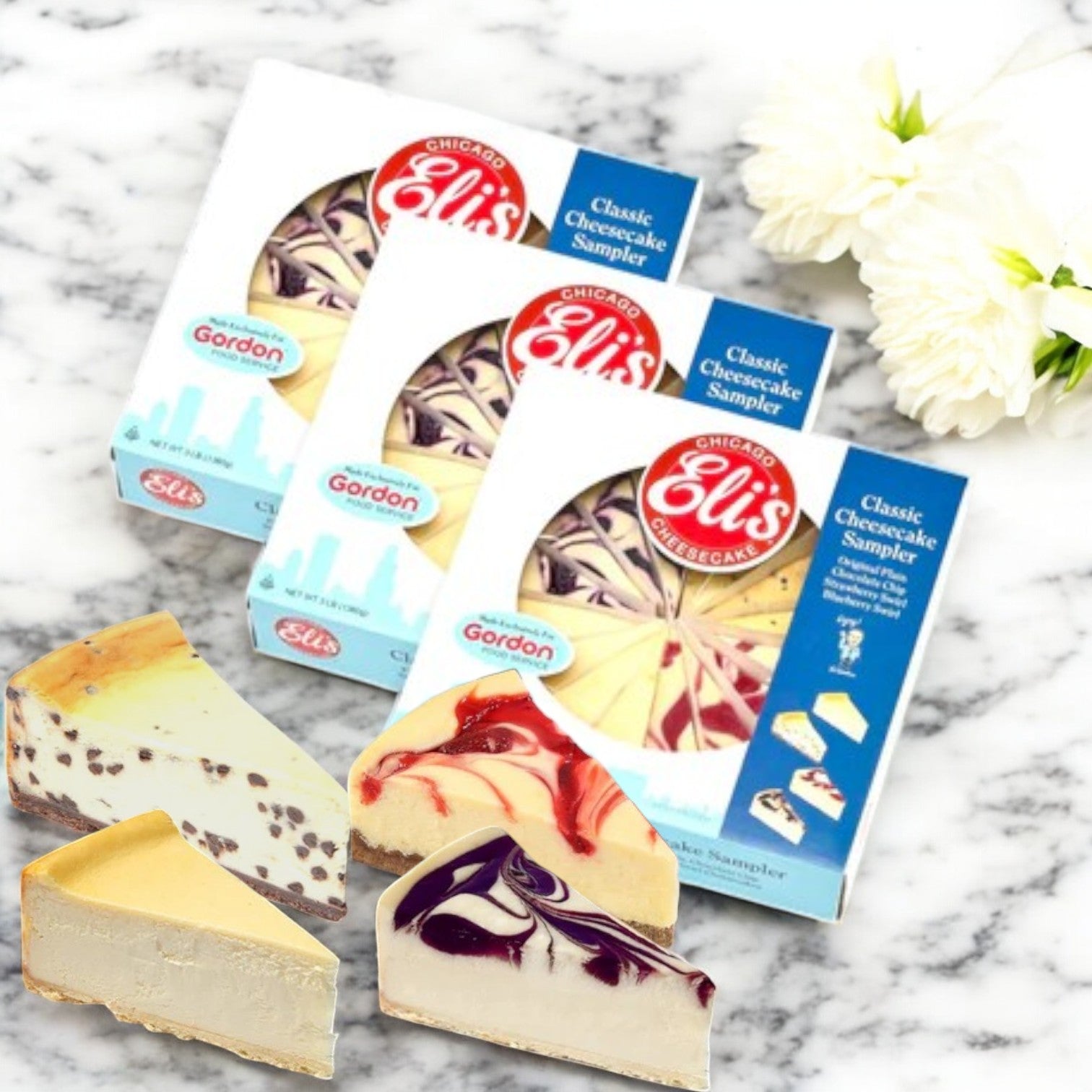 Three boxes of Eli's 9" Classic Cheesecake Sampler - 3 Count from Eli's Cheesecake sit elegantly on a marble surface, featuring a variety of slices. Delicate white flowers enhance this delightful display.
