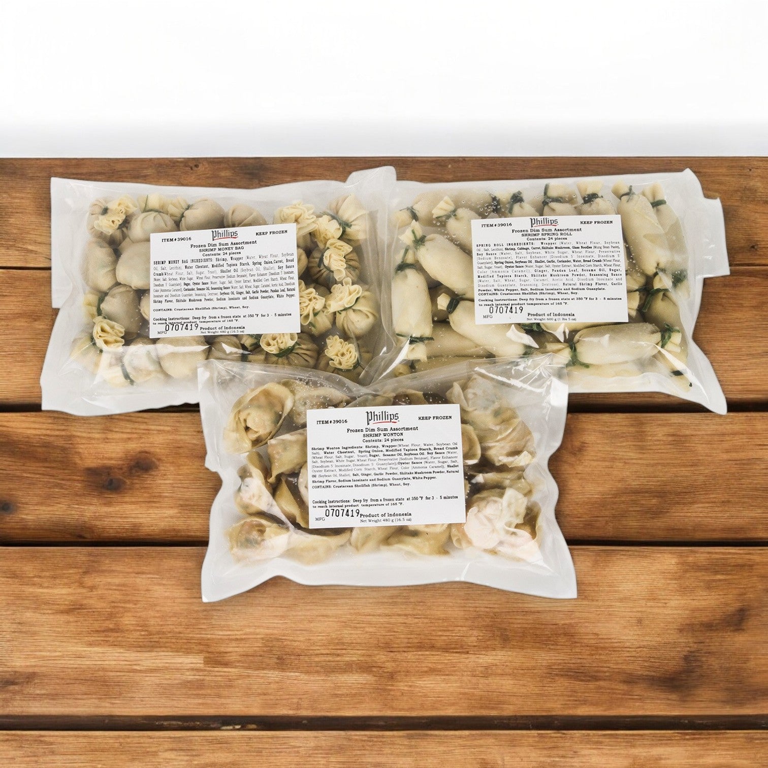 Three bags of frozen Phillips Foods appetizers, specifically dim sum fried pieces, are wrapped in clear plastic and placed on a wooden surface.