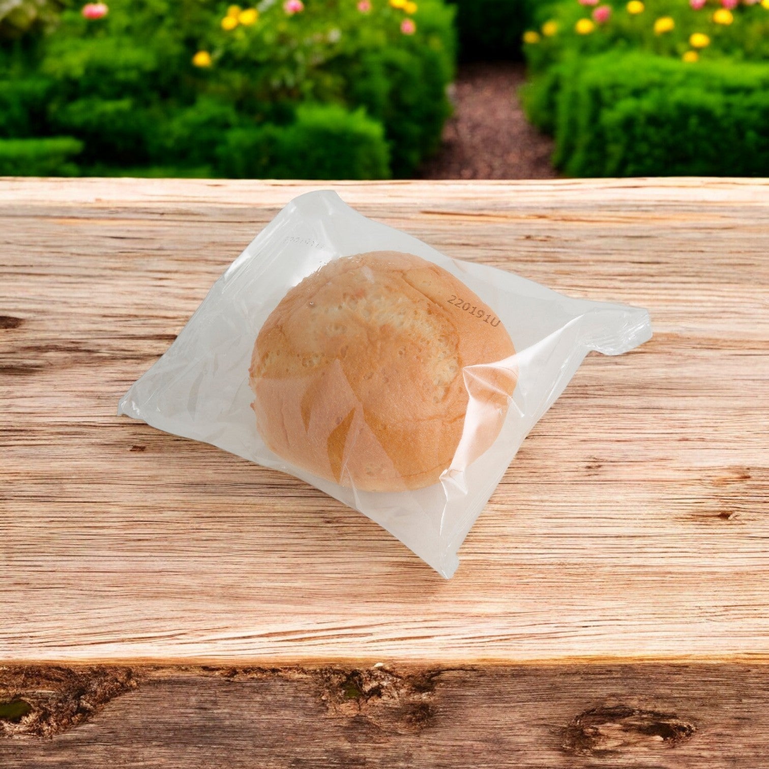 Udi's Bun Hamburger Gluten Free, a soft and sturdy 4-inch roll from Udi's, is individually wrapped in clear plastic packaging and rests on a wooden surface with a garden view.