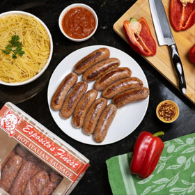Plate of cooked Esposito's Finest Quality Hot Italian Sausage surrounded by uncooked pasta, a bowl of marinara sauce, sliced red peppers, crushed red pepper flakes, and packaged sausages on a dark table. An authentic Italian recipe crafted with premium ingredients for a delightful dining experience.