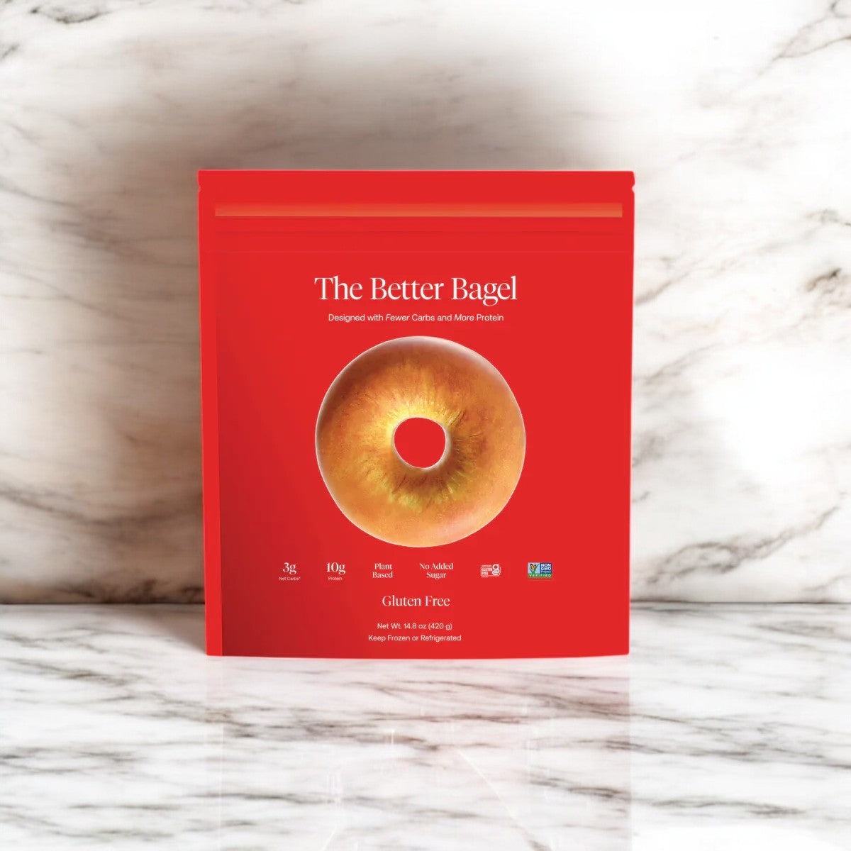 The eye-catching red packaging of BetterBrand's "The Gluten-Free Better Bagel" features a tempting bagel image and proclaims it as high-protein, gluten-free, and low-carb, all elegantly displayed on a sleek marble surface.