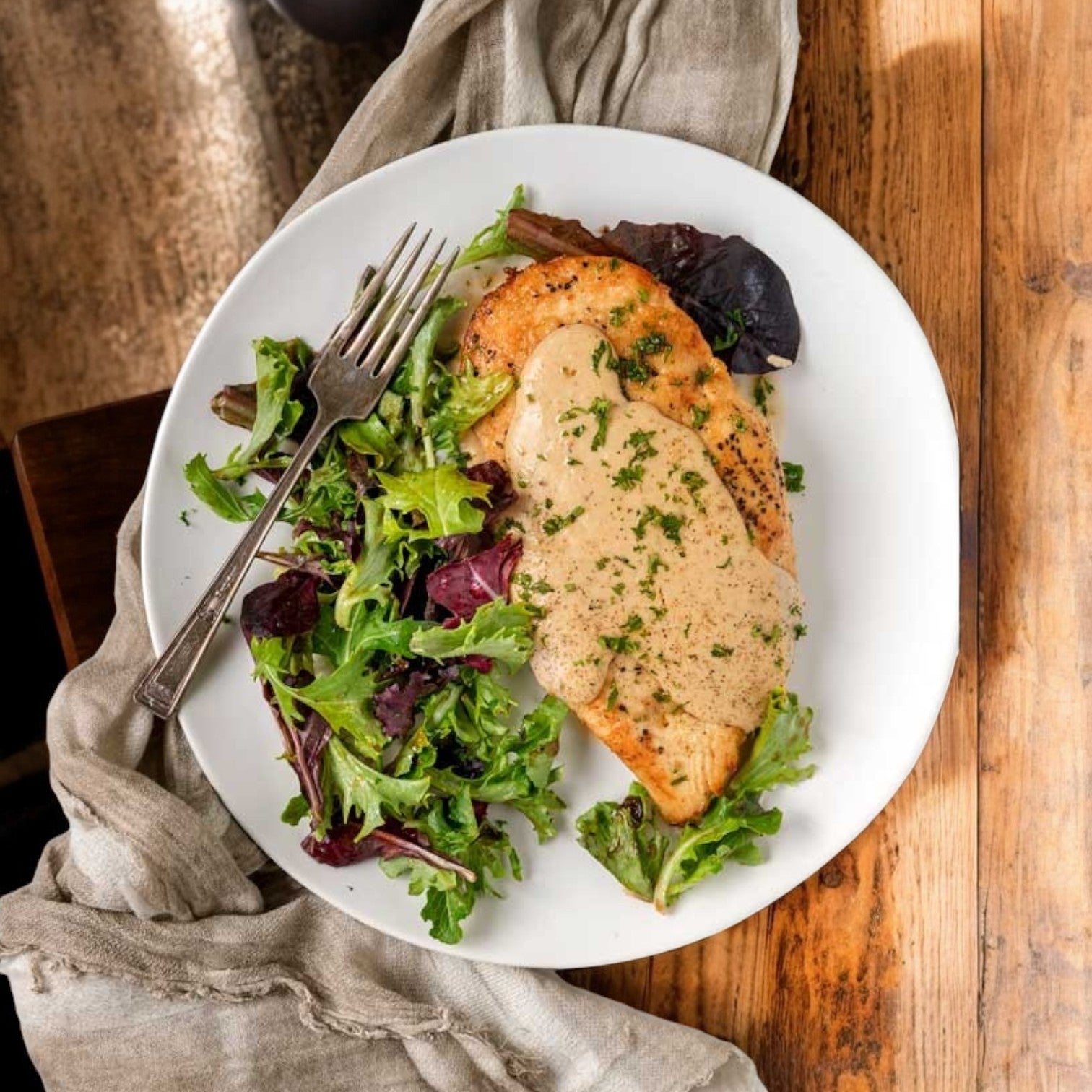 A plate of Garden Valley Grilled Halal Chicken Breast Fillets, fully cooked and topped with creamy sauce, rests on a wooden table next to mixed greens, accompanied by a cloth napkin and fork.