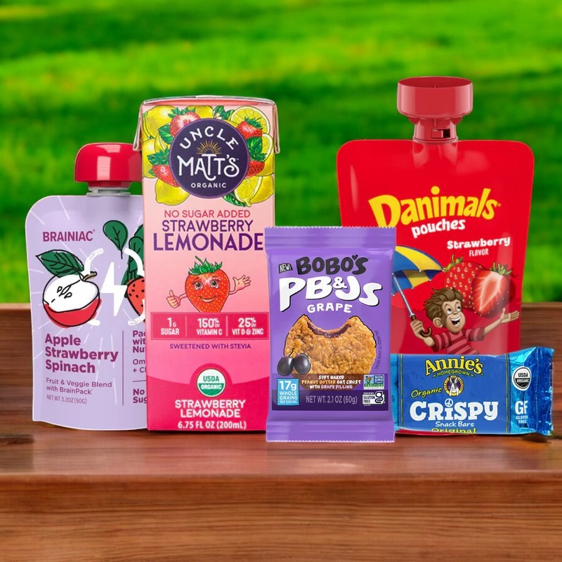 An assortment of children's snacks from Easy Lunches, including Gluten Free Lunch! 5 Total Items, is displayed on a wooden table with organic strawberry lemonade, apple strawberry spinach juice, a PB&J bar, and a gluten-free crispy snack bar against a grassy backdrop.