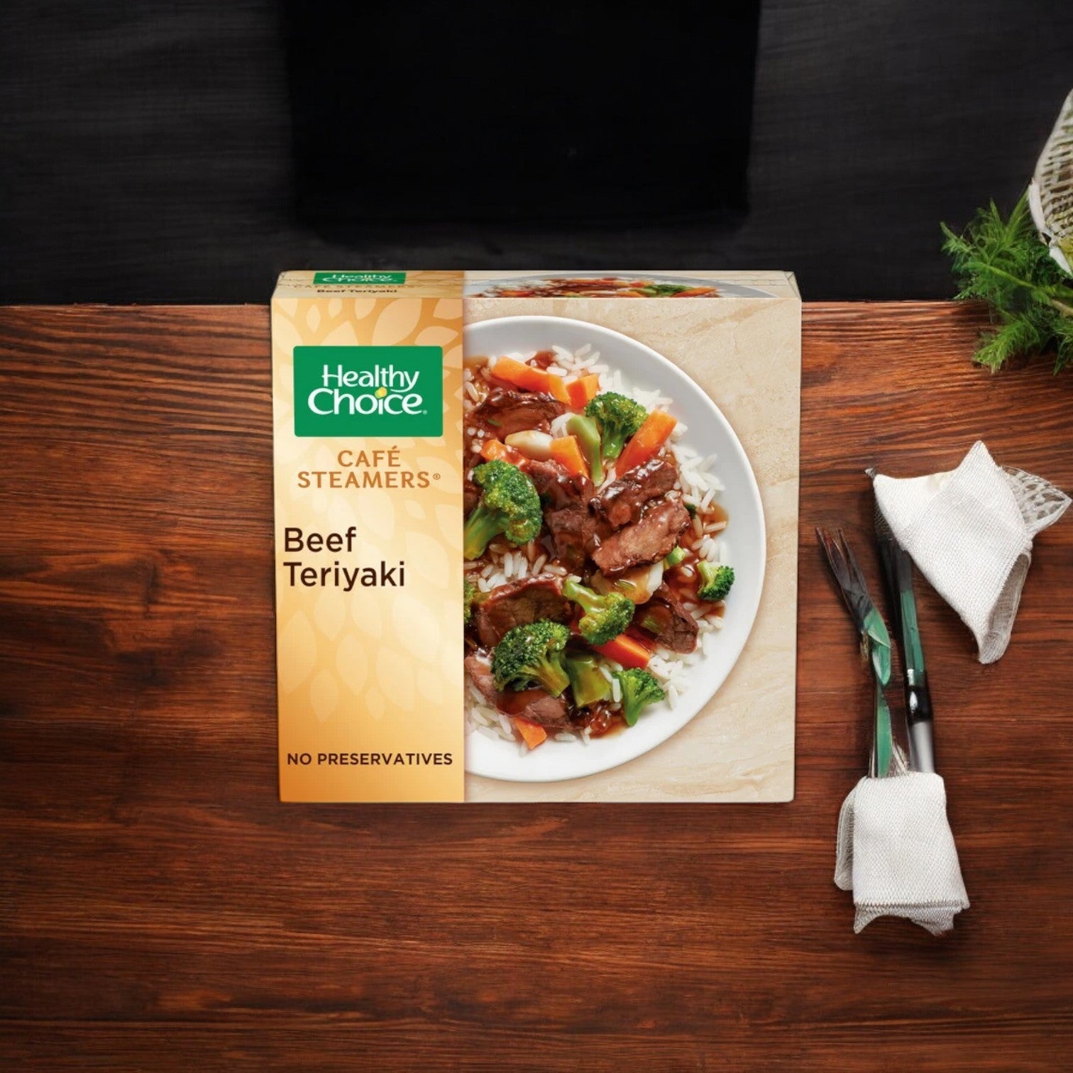 A Healthy Choice Café Stmr Beef Teriyaki, 9.5 oz. - 1 Count, package is enticingly displayed on a wooden table, highlighting the innovative steam-cooking technology from the brand Healthy Choice. Beside it, a set of black utensils elegantly wrapped in white napkins awaits your dining pleasure.