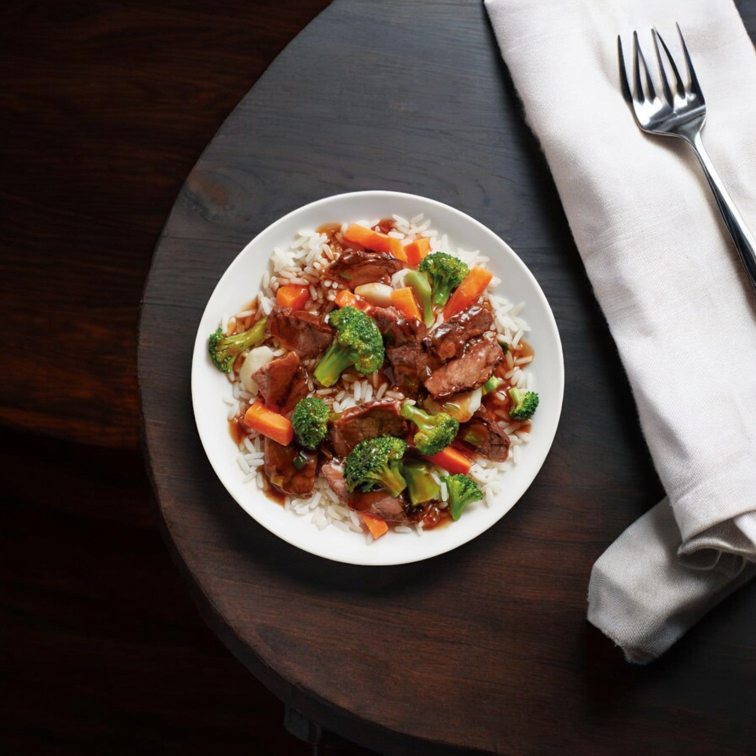 A serving of Healthy Choice Café Steamer Beef Teriyaki, highlighting the advantages of steam-cooking technology, is garnished with broccoli and carrots next to a folded white napkin and fork on a dark wooden table.