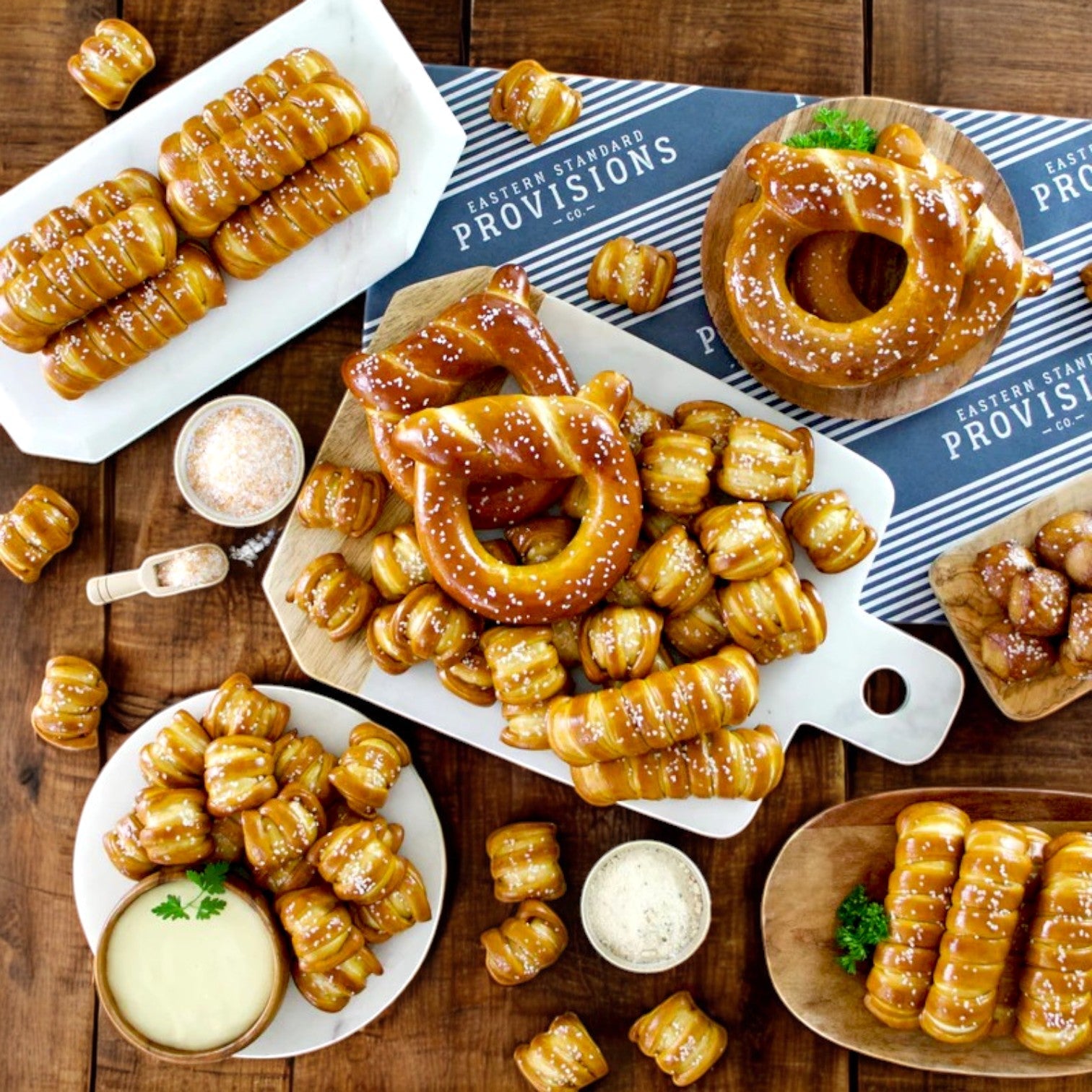 Assorted artisanal soft pretzels in various shapes and salted toppings are on a wooden table with dipping sauces. A cloth displays "Eastern Standard Provisions Pretzel 3-Pack Combo," highlighting favorites like the three cheese classic.