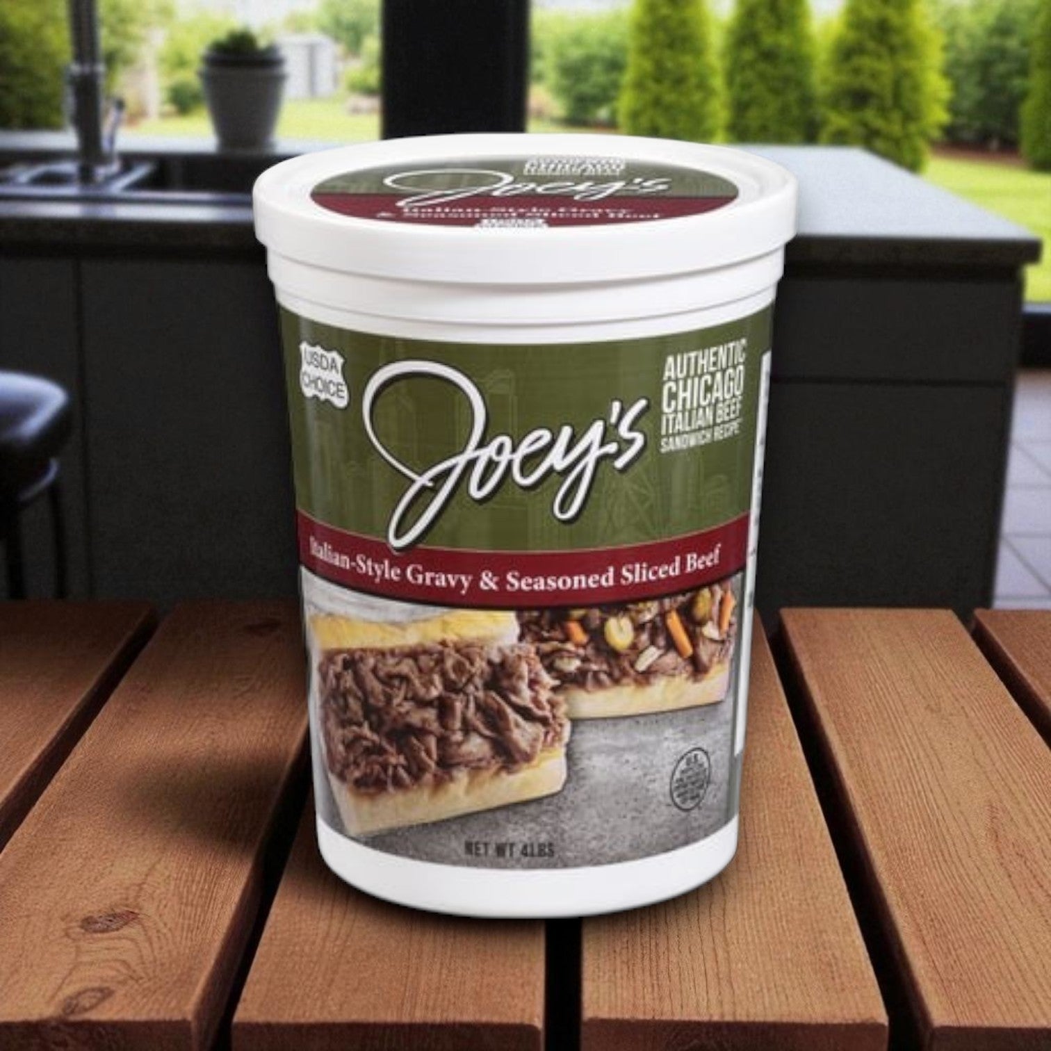 A container of Endless Market’s Joey’s Italian-Style Sliced Beef & Vienna Beef Giardiniera Combo sits on a wooden table—ideal for classic Chicago Italian beef sandwiches—with a kitchen and outdoor greenery in the background.