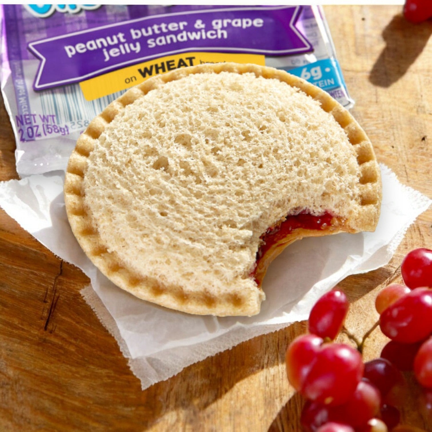An Uncrustable Smuckers Peanut Butter & Grape Jelly sandwich, with one bite taken out, is placed on a white napkin next to a bundle of grapes and its packaging—ideal for convenient snacking. This deliciously easy treat comes from the 10 Count - 2 Box pack, providing a total of 20 sandwiches for your enjoyment.