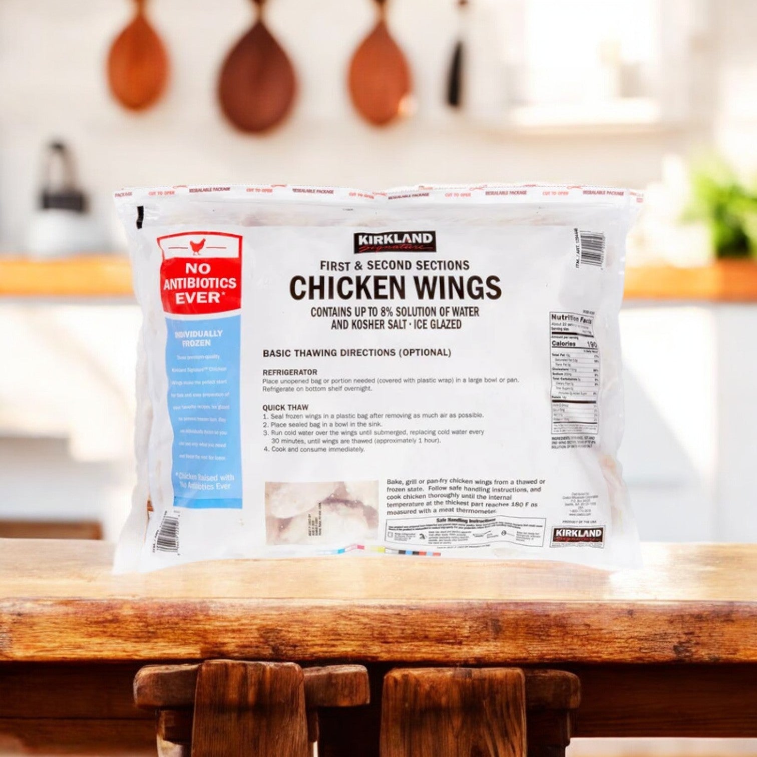A package of Kirkland Signature Chicken Wings (10 lbs., 3 bags, 30 lbs. total) rests on a wooden surface, labeled "No Antibiotics Ever" with thawing instructions. Perfect for versatile cooking uses, blurred kitchen shelves and utensils form the backdrop.