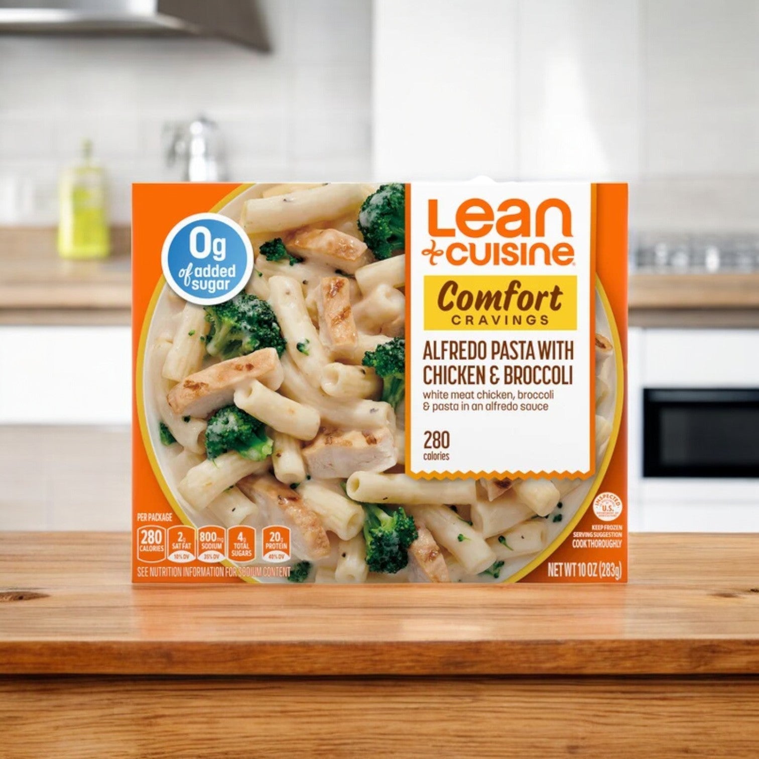 A box of Lean Cuisine Alfredo Pasta with Chicken & Broccoli, 10 oz. - 1 Count, sits on a kitchen counter. The packaging highlights 0g of added sugar and 280 calories per serving, making it a perfect frozen entrée choice.