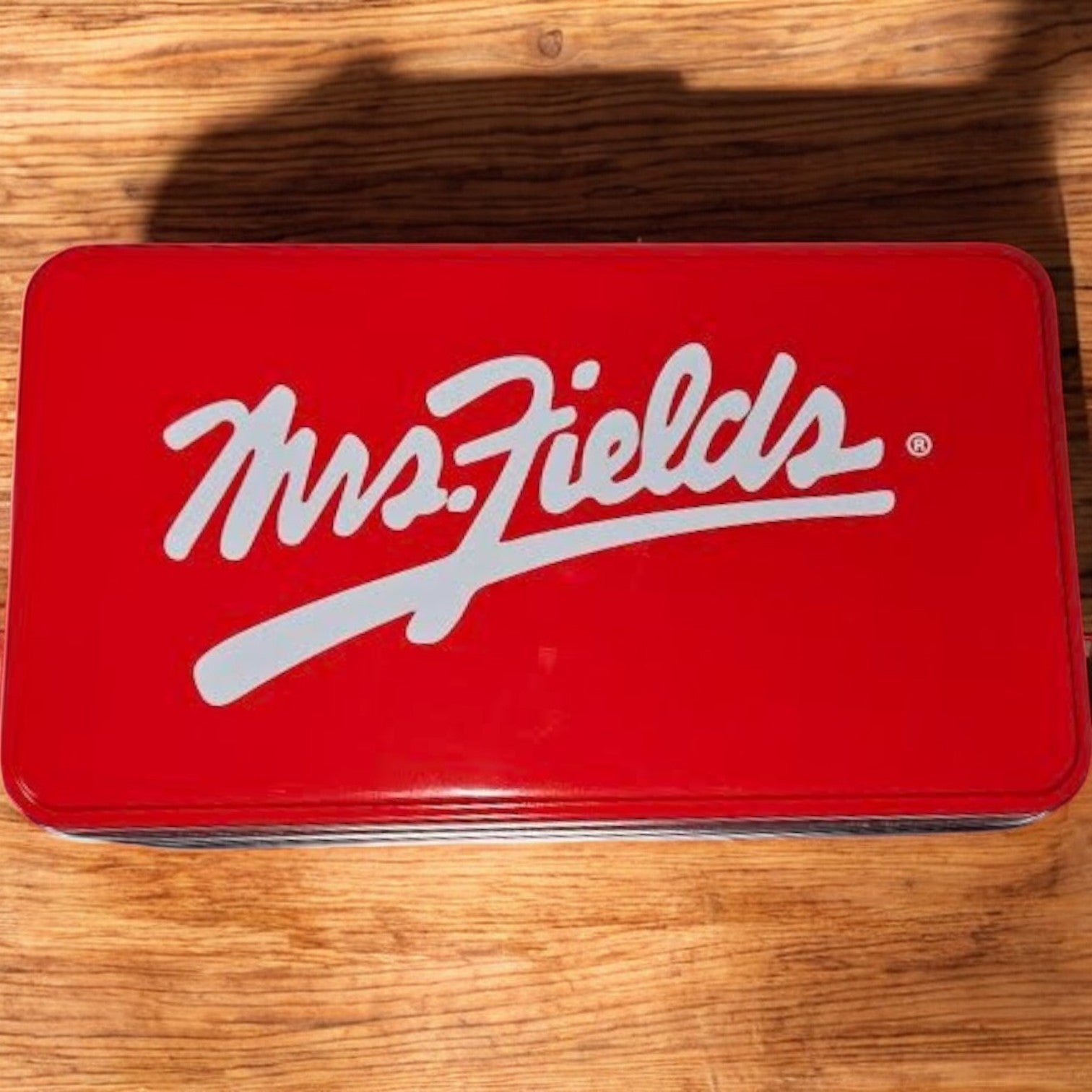 A Mrs. Fields Signature Tin, with a red rectangular design and elegant white script logo, rests on a wooden surface, filled with 24 Nibblers Bite-Sized Assorted Cookies for those who cherish these delights.