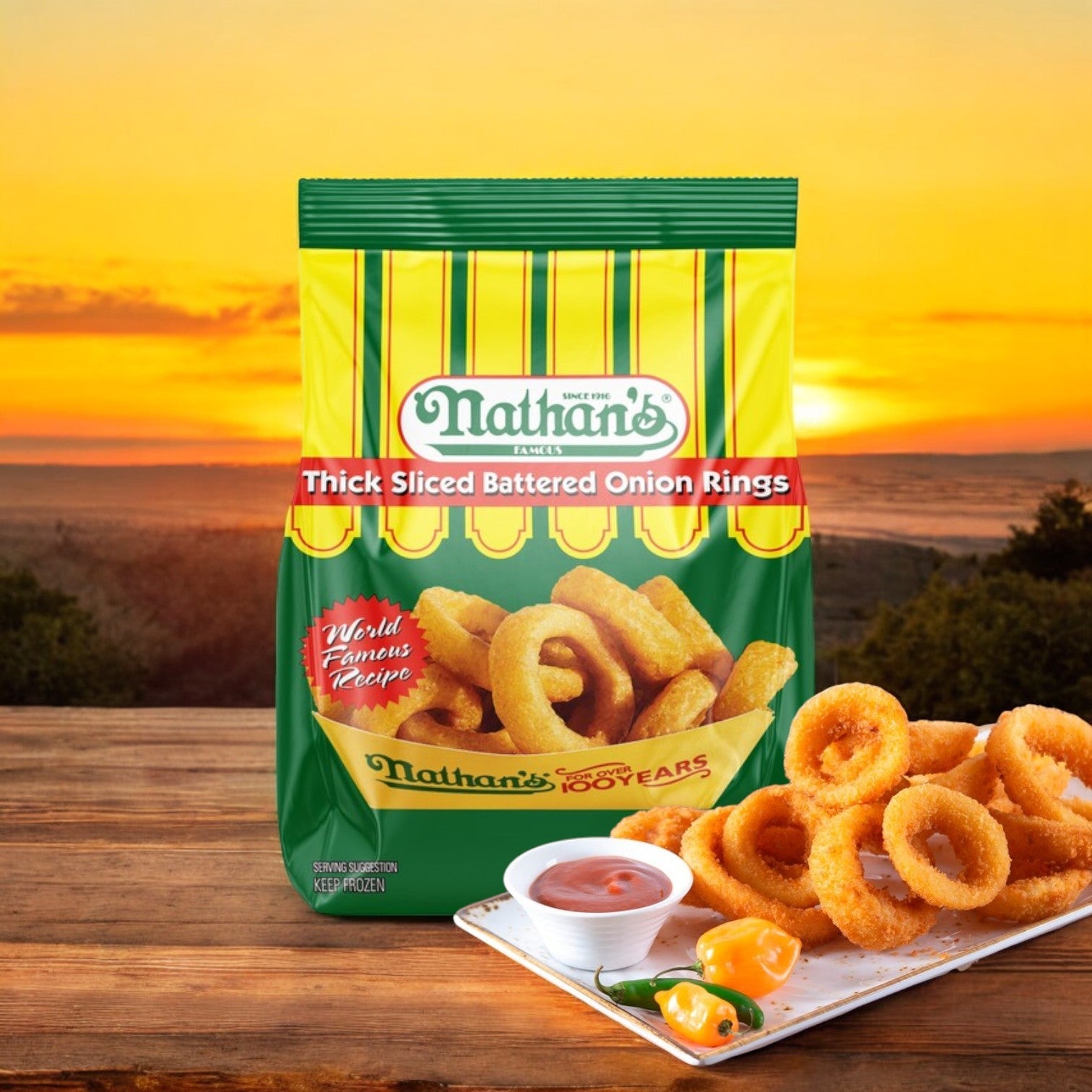 A 16 oz package of Nathan’s Famous Battered Thick-Sliced Onion Rings graces the table, accompanied by dipping sauce and a vibrant yellow pepper, all set against a stunning sunset backdrop.