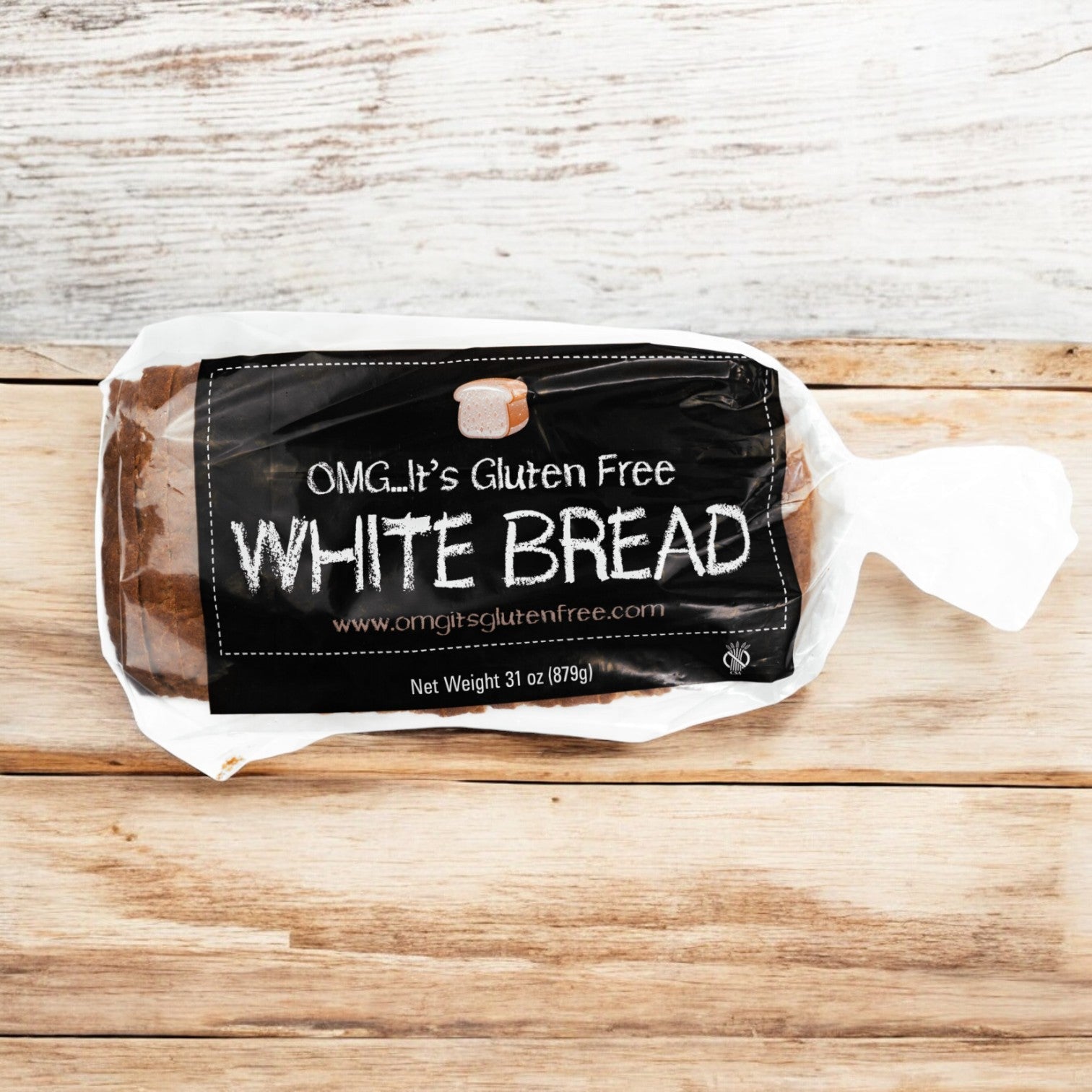 A loaf of OMG It's Gluten Free, White Bread Loaf (31 oz.) in clear packaging with a black label lies on a wooden surface. The label reads "OMG. It's Gluten Free WHITE BREAD." Alongside, there are individually sized, soft and fluffy Gluten-Free Dinner Rolls.