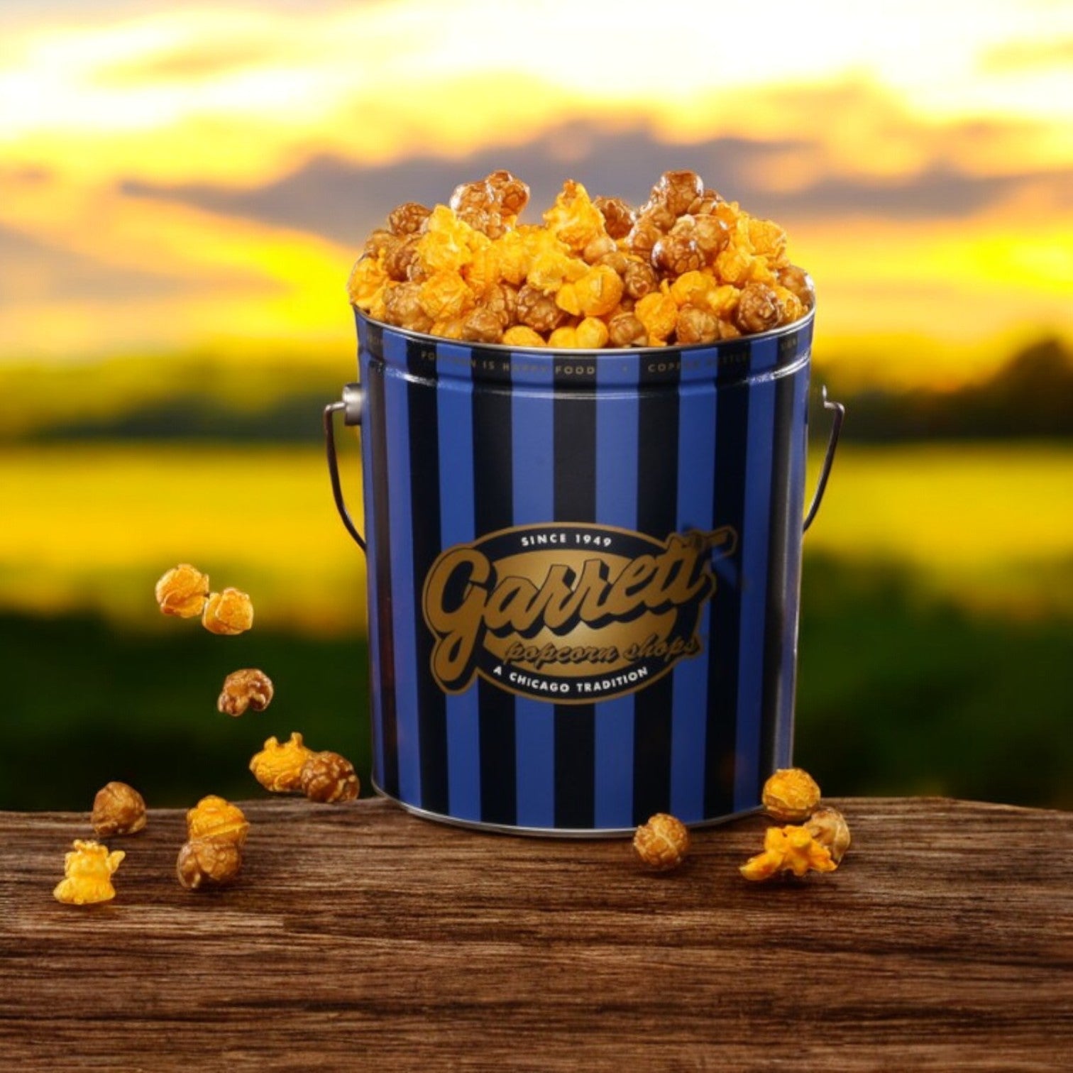 A classic tin of Garrett Popcorn - Garrett Mix by Garrett rests on a wooden surface, filled with CaramelCrisp popcorn spilling out against a blurred field backdrop.