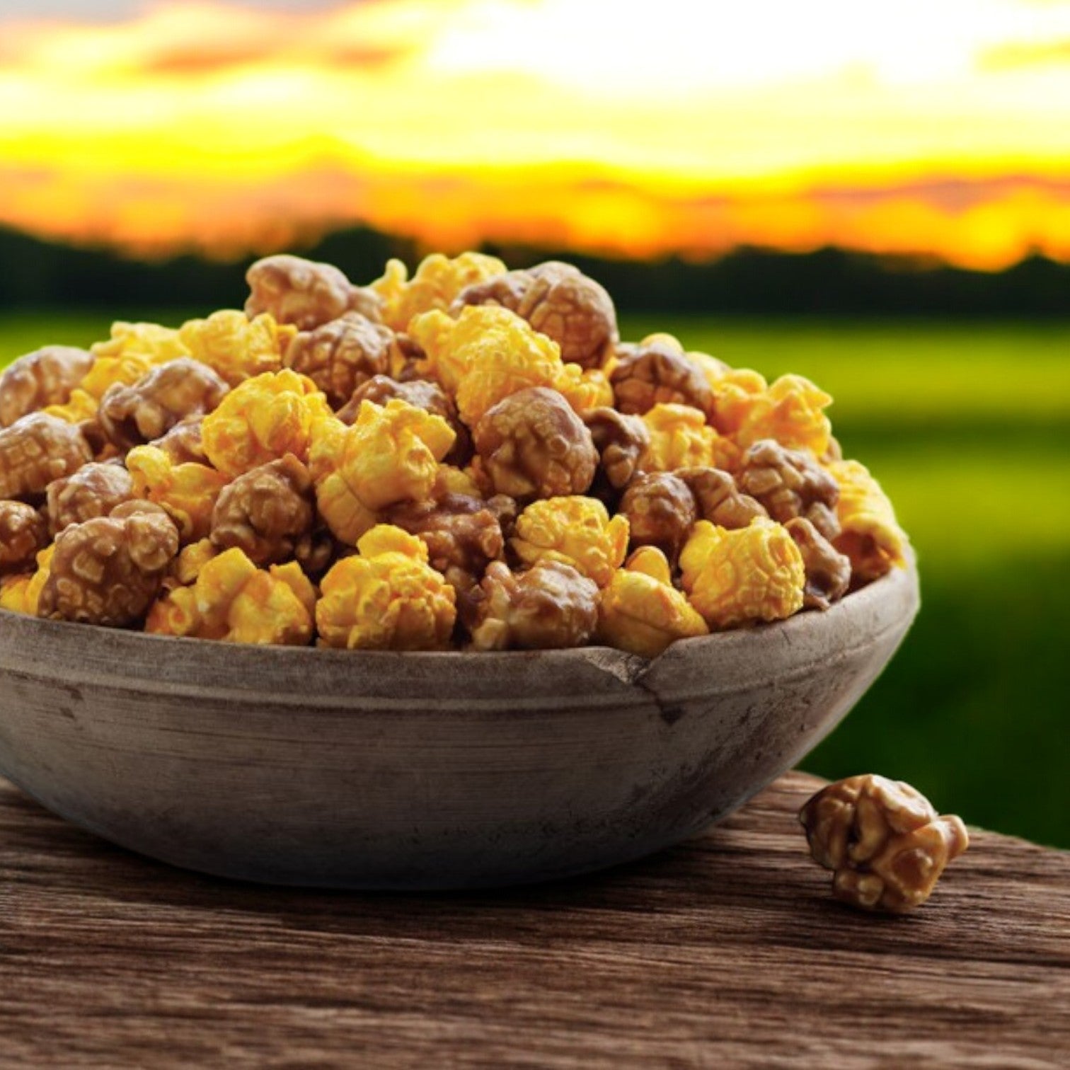 A classic tin of Garrett Popcorn's Garrett Mix, featuring CaramelCrisp and CheeseCorn, is placed on a wooden surface against the backdrop of a blurred sunset and field.