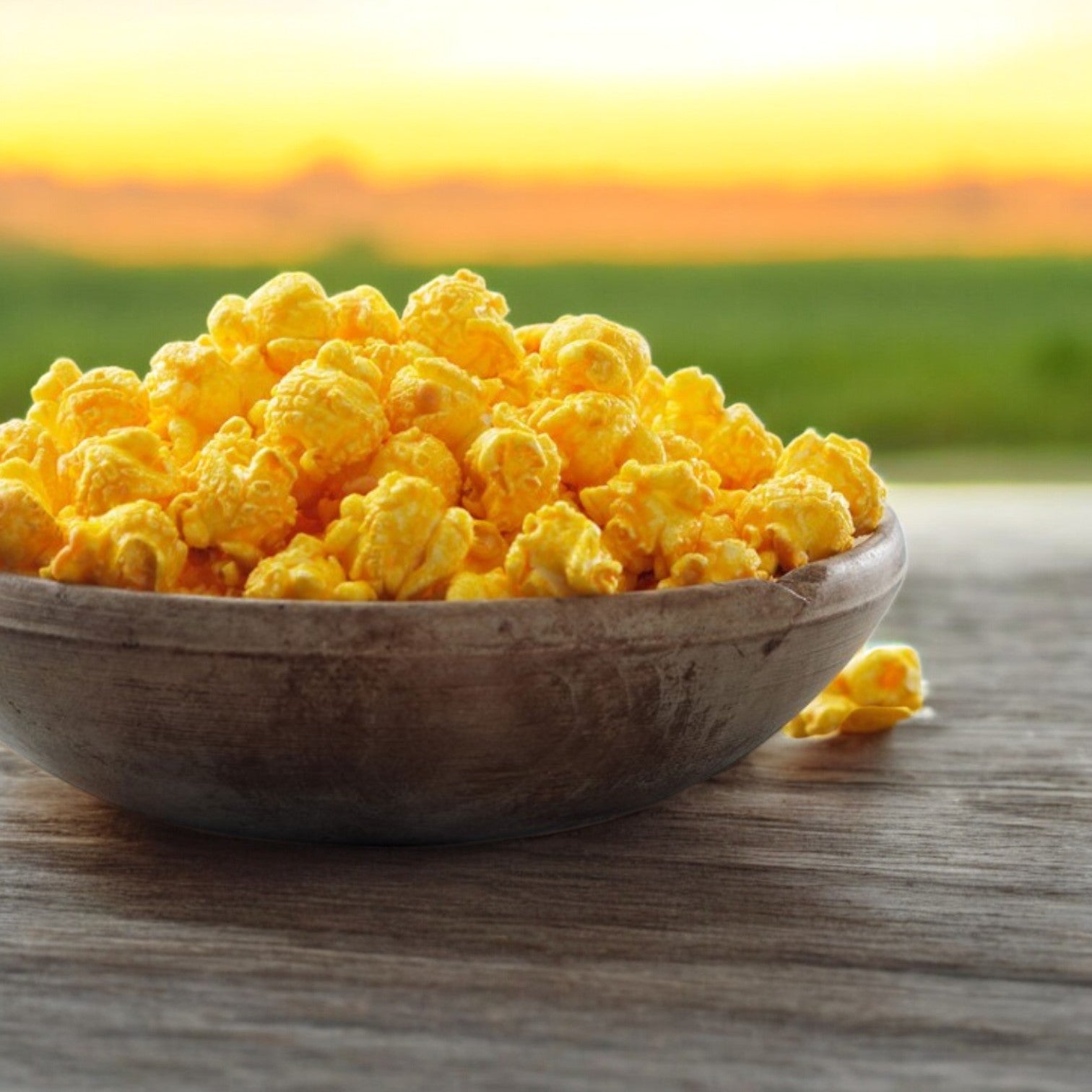 A wooden bowl filled with Garrett Popcorn's Cheese Corn, 14 Cups in a Classic Tin Size, rests on a wooden surface with the blurred backdrop of a sunset over lush green fields.