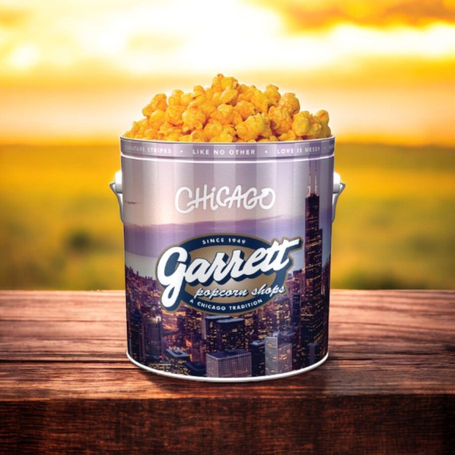 A Garrett Popcorn Classic Tin of Cheese Corn, featuring 14 cups, sits on a wooden surface with its vivid popcorn contrasting beautifully against the soft sunset backdrop.
