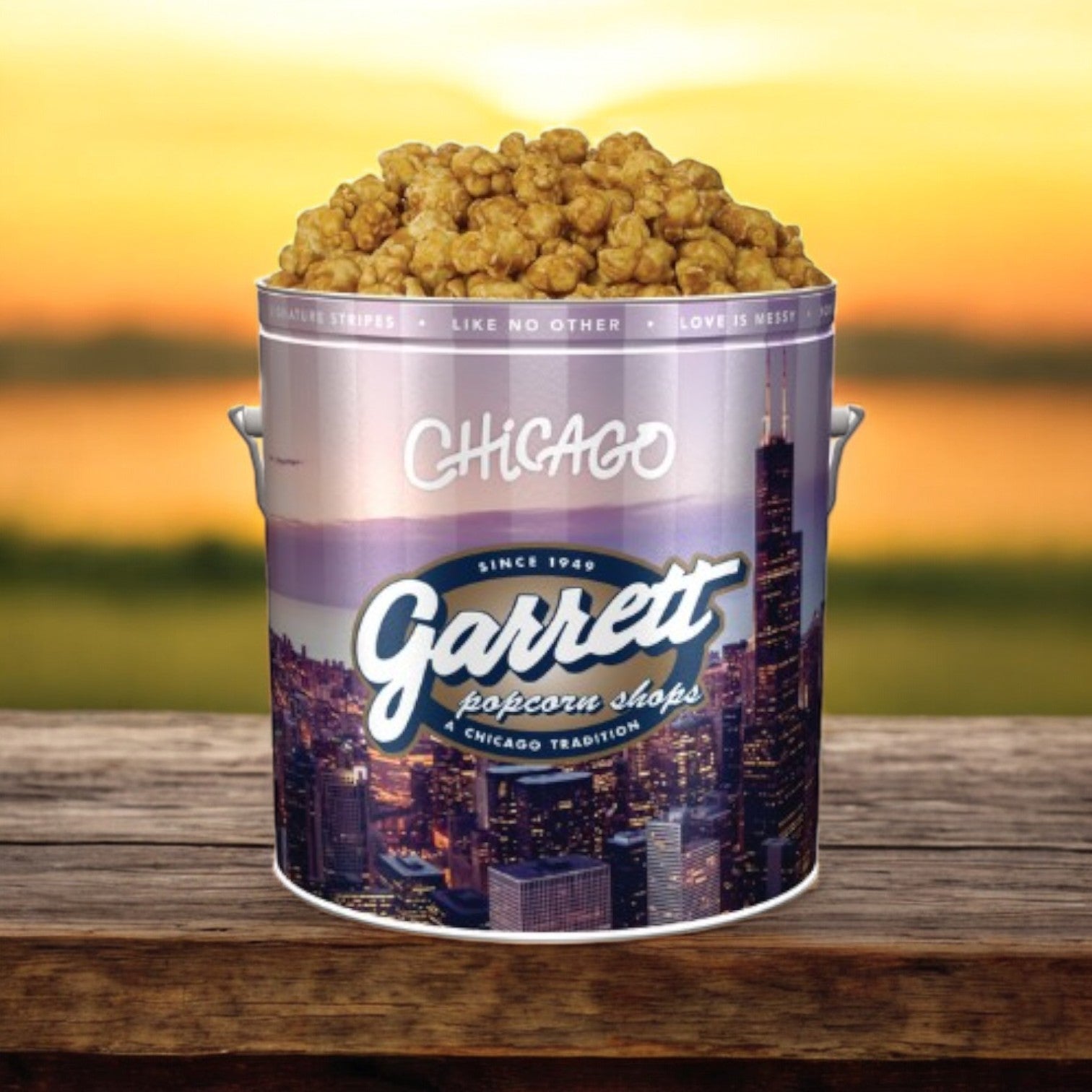 A Classic Tin of Garrett Popcorn - Caramel Crisp, containing 14 cups, is placed on a wooden surface against a stunning sunset backdrop.