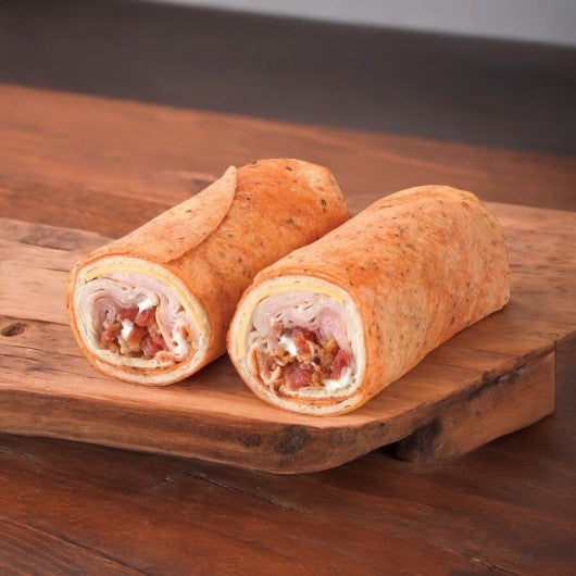 Two E.A. Sween Turkey Club Wraps, each featuring turkey breast, cheddar cheese, vegetables, crispy bacon, ham, and ranch dressing peek through layers on a wooden board. Ships frozen with a 30-day shelf life out of the freezer!.