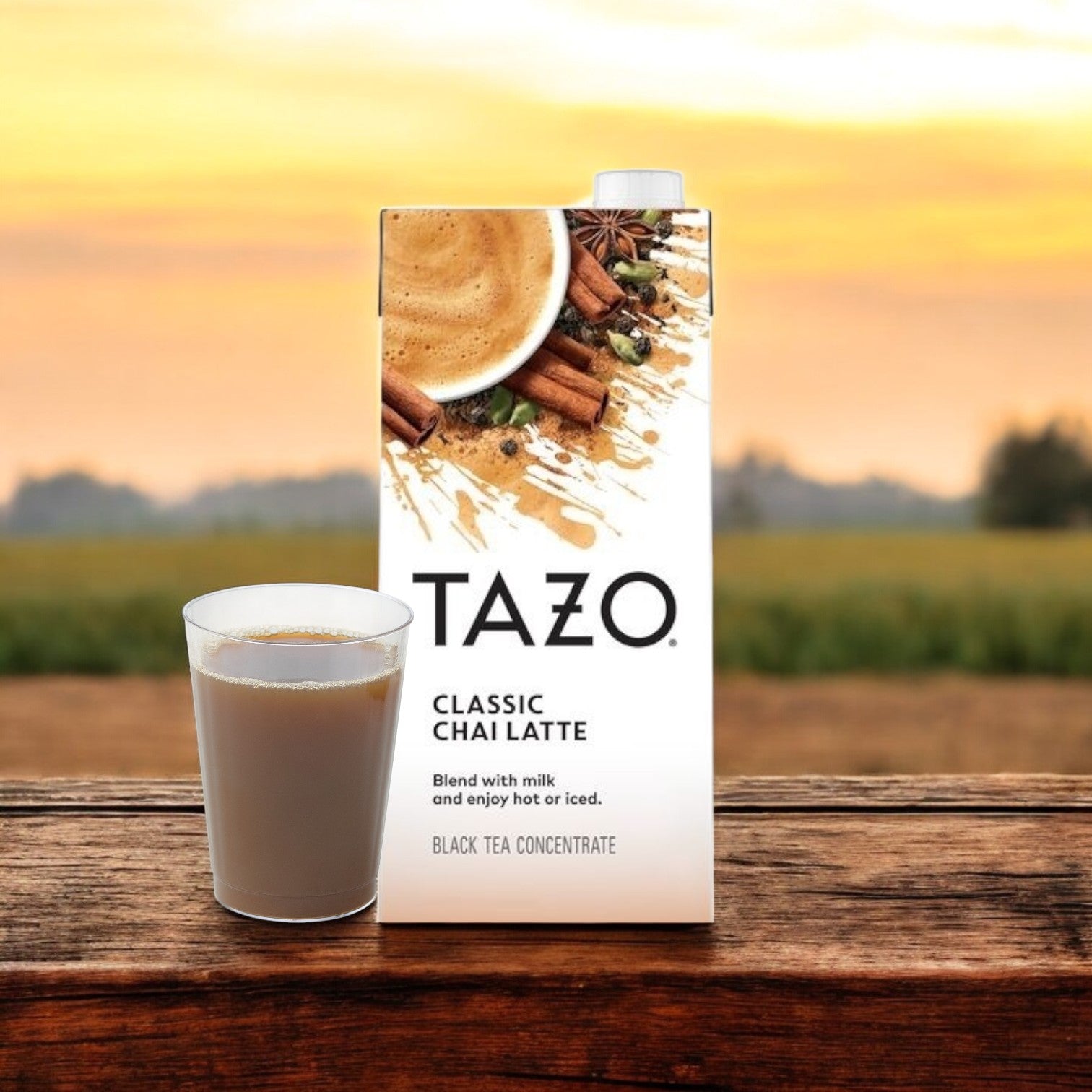 A Tazo Tea Chai Classic Concentrate carton and a filled glass sit elegantly on a wooden surface, framed by a sunset over a field. The rich aroma of this black tea blend enhances the tranquil evening scene.