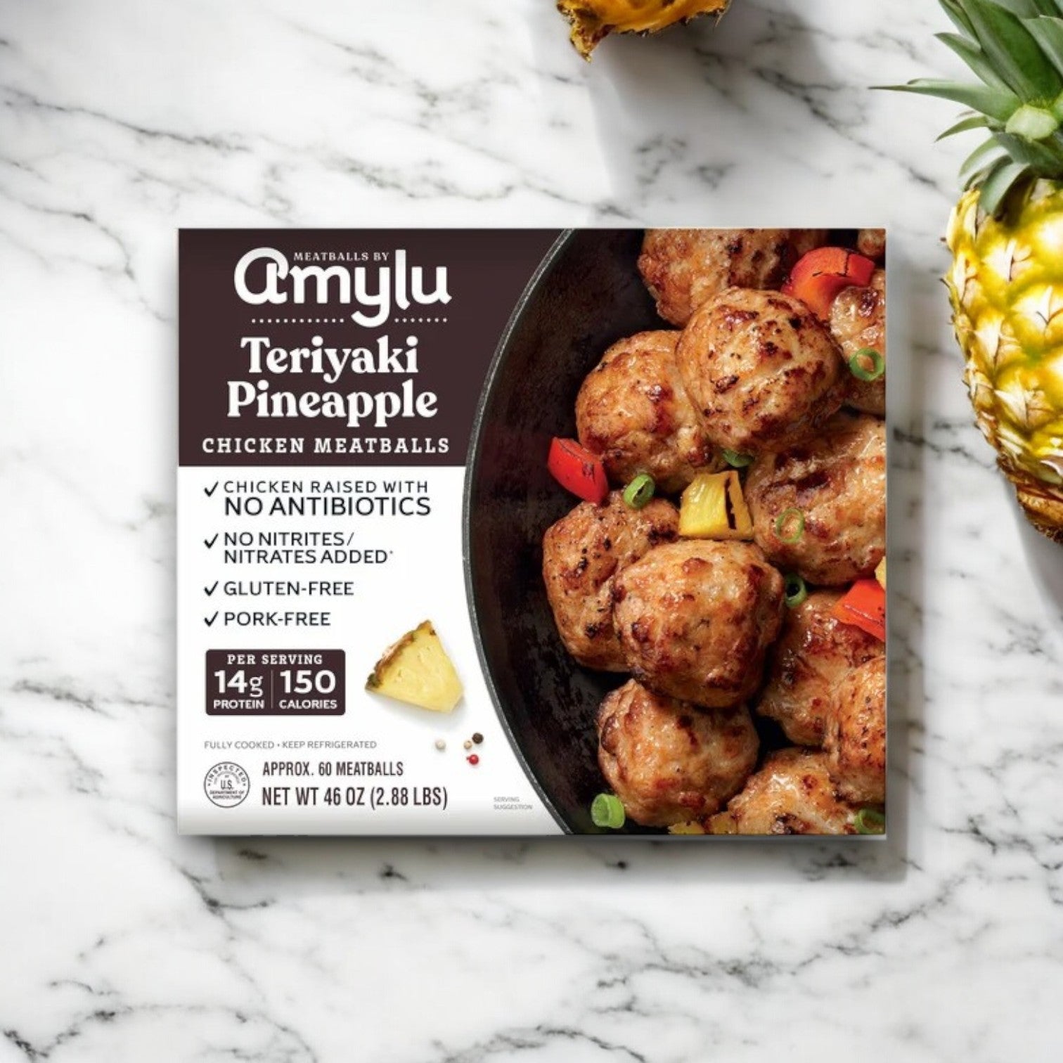 On a marble surface, the Amylu Teriyaki Pineapple Chicken Meatballs packaging highlights its premium ingredients such as gluten-free benefits and antibiotic-free chicken. An enticing image shows the meatballs with pineapple, promising a delicious experience.