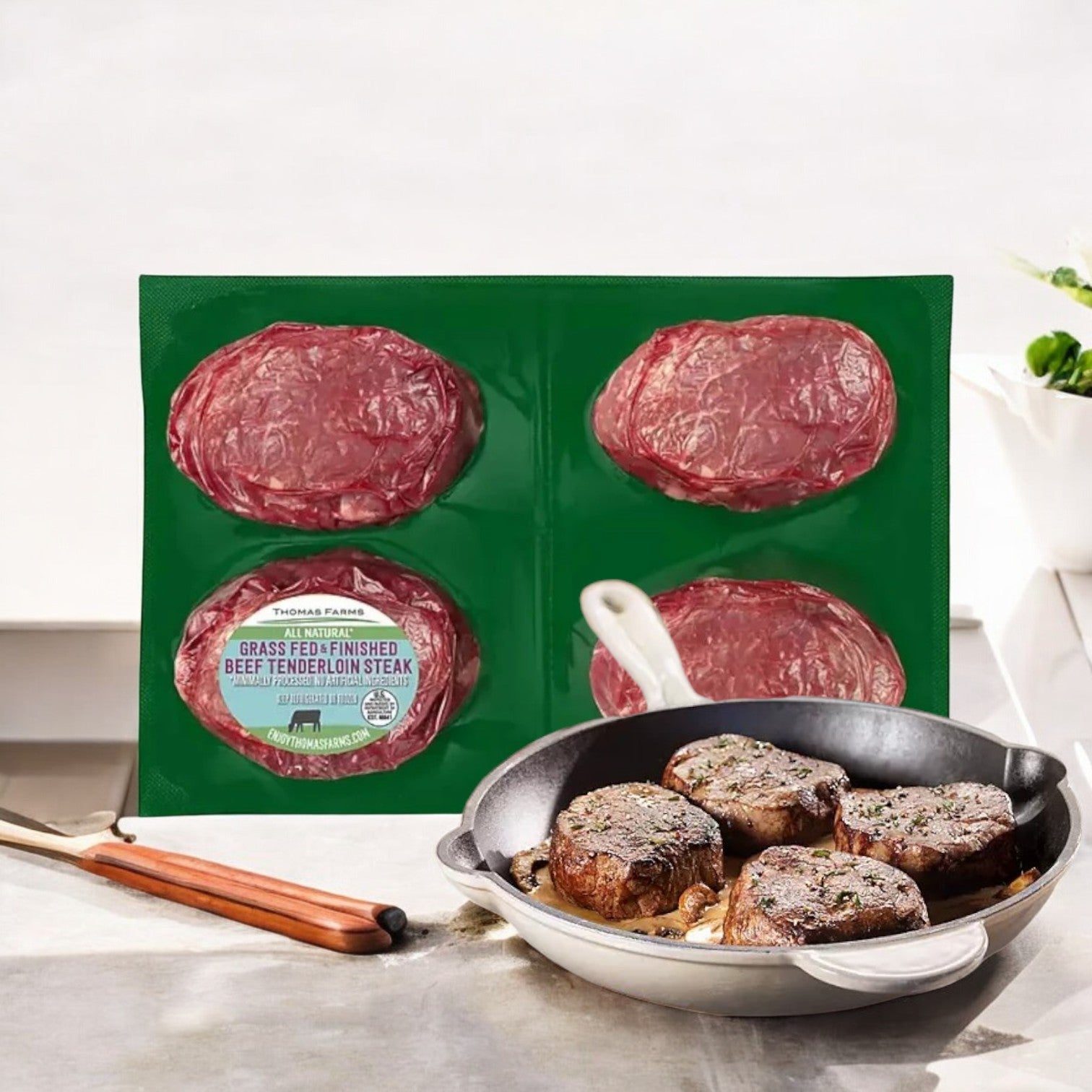A package of premium-quality, grass-fed beef tenderloin steaks sits beside a skillet on the kitchen counter containing perfectly cooked Thomas Farms New York Strip Steak from their Grass Fed Beef line, featuring 1 pack with 2 steaks.