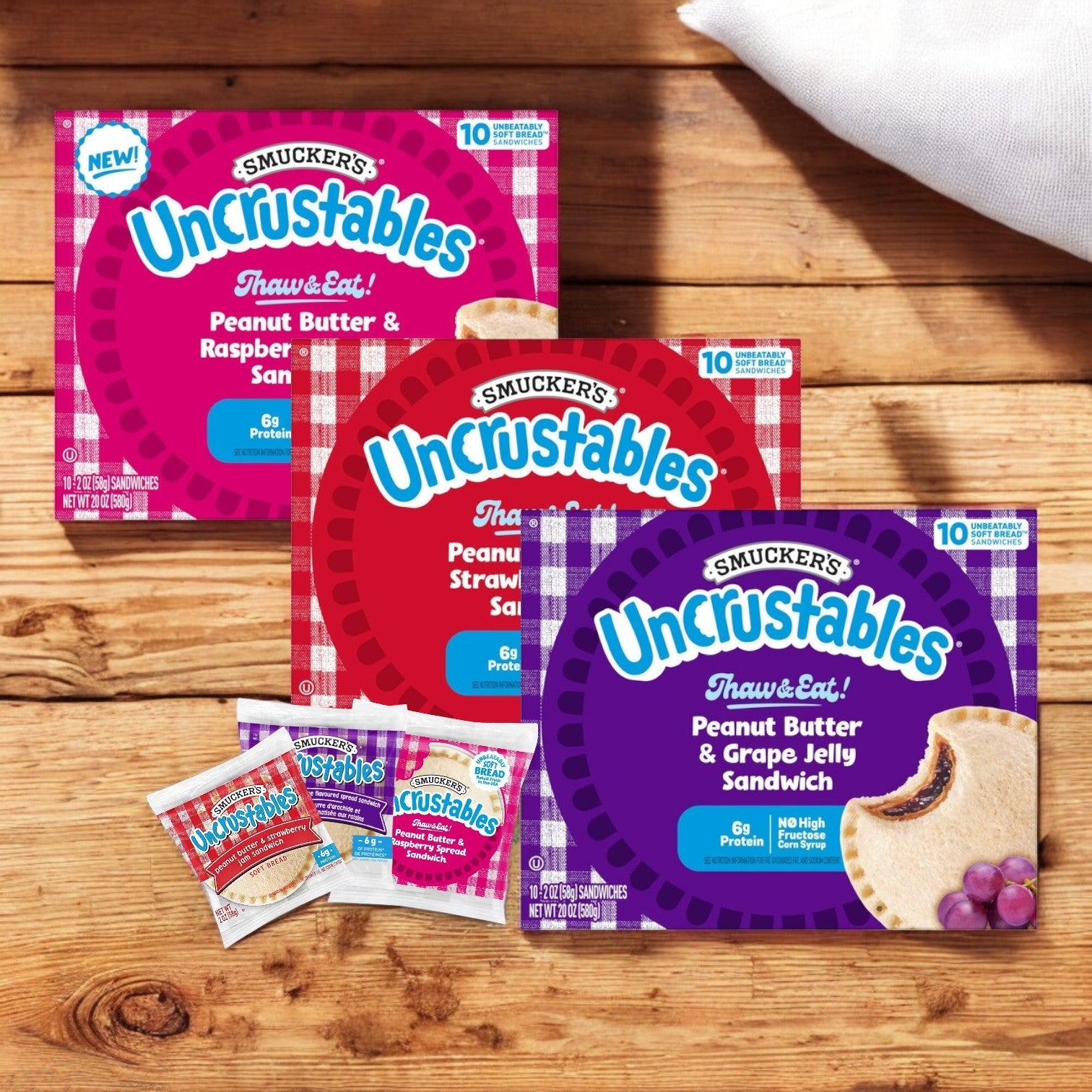 Three boxes of Uncrustables Combo of 3 – 10 Count Each by Uncrustable Smuckers rest on a wooden surface, featuring the delightful Variety Pack with Peanut Butter & Raspberry, Peanut Butter & Strawberry, and Peanut Butter & Grape Jelly flavors—all on soft crustless bread.