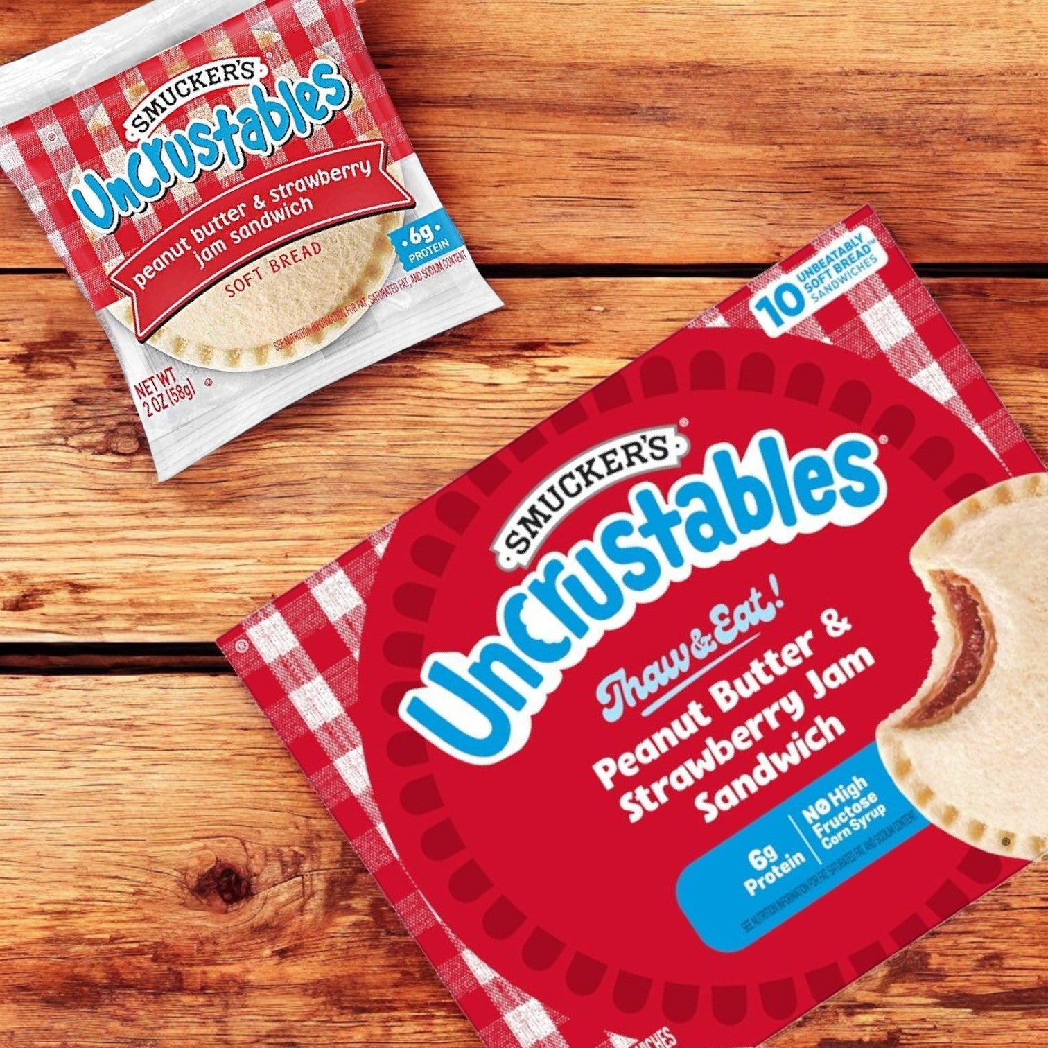 Two Uncrustable Smuckers packages sit on a wooden surface: one individually wrapped peanut butter and strawberry jam sandwich, and an Uncrustables Combo of 3 – 10 Count Each box containing ten crustless sandwiches.
