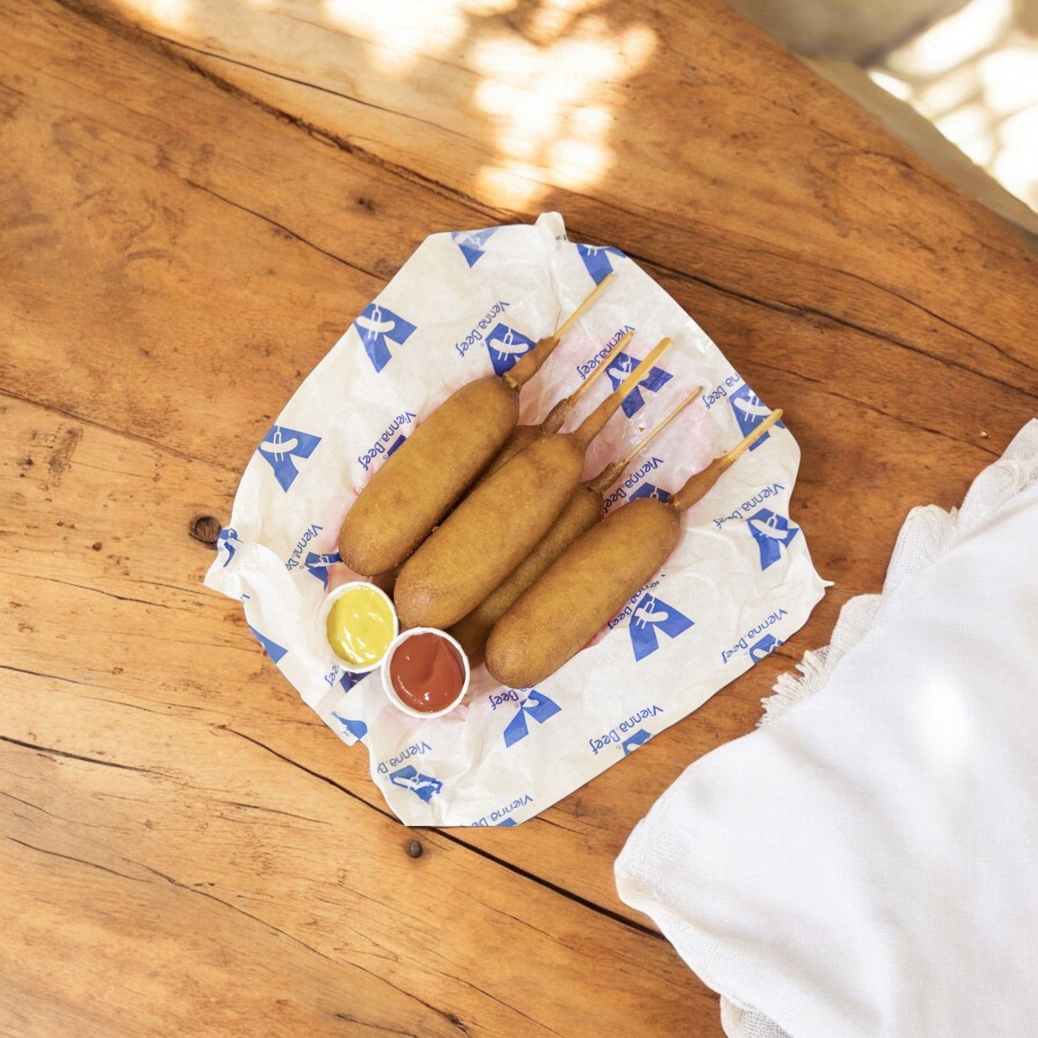 Three Vienna Beef Corn Dogs (3.25 oz.) from a 24-count pack rest on a paper-lined tray with mustard and ketchup on a wooden table, encased in golden cornmeal batter.