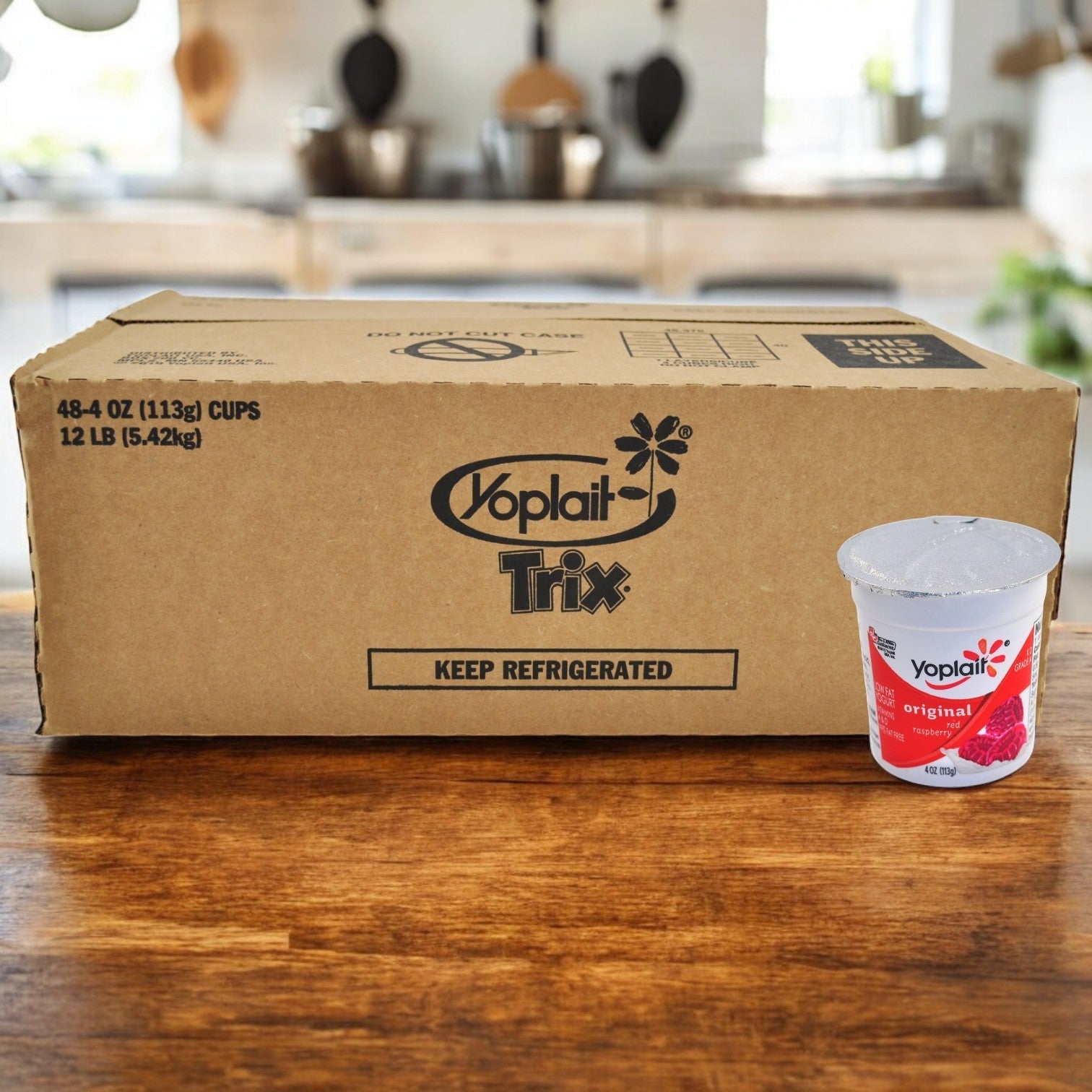 A big box marked "Yoplait Original Raspberry and Peach" is on a wooden table, beside a small container of Yoplait original strawberry yogurt, famous for its creamy texture and real fruit flavors.