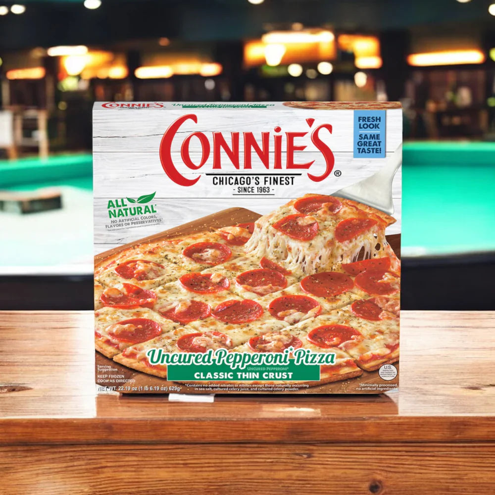 A box of Easy Lunches' Connie's Bundle! 4 Total Items, with uncured pepperoni and classic thin crust, rests on a wooden surface against a blurred pool table background.