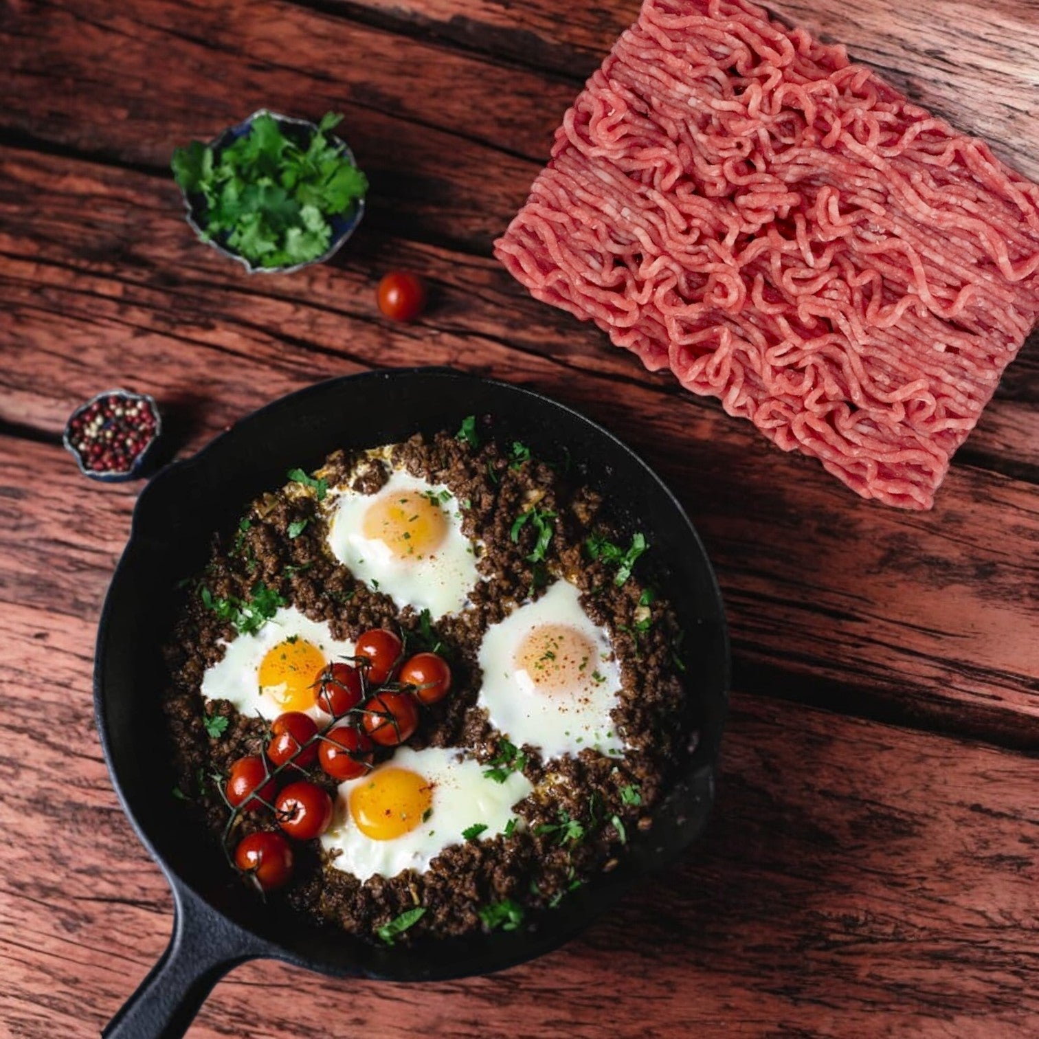 A cast iron skillet sizzles with three eggs, Australian lamb, and cherry tomatoes. Beside the skillet on the wooden table lie a bunch of parsley, ground pepper, and Patuxent Farms Halal Ground Lamb 80/20 from Australia in a 296 oz vacuum package.