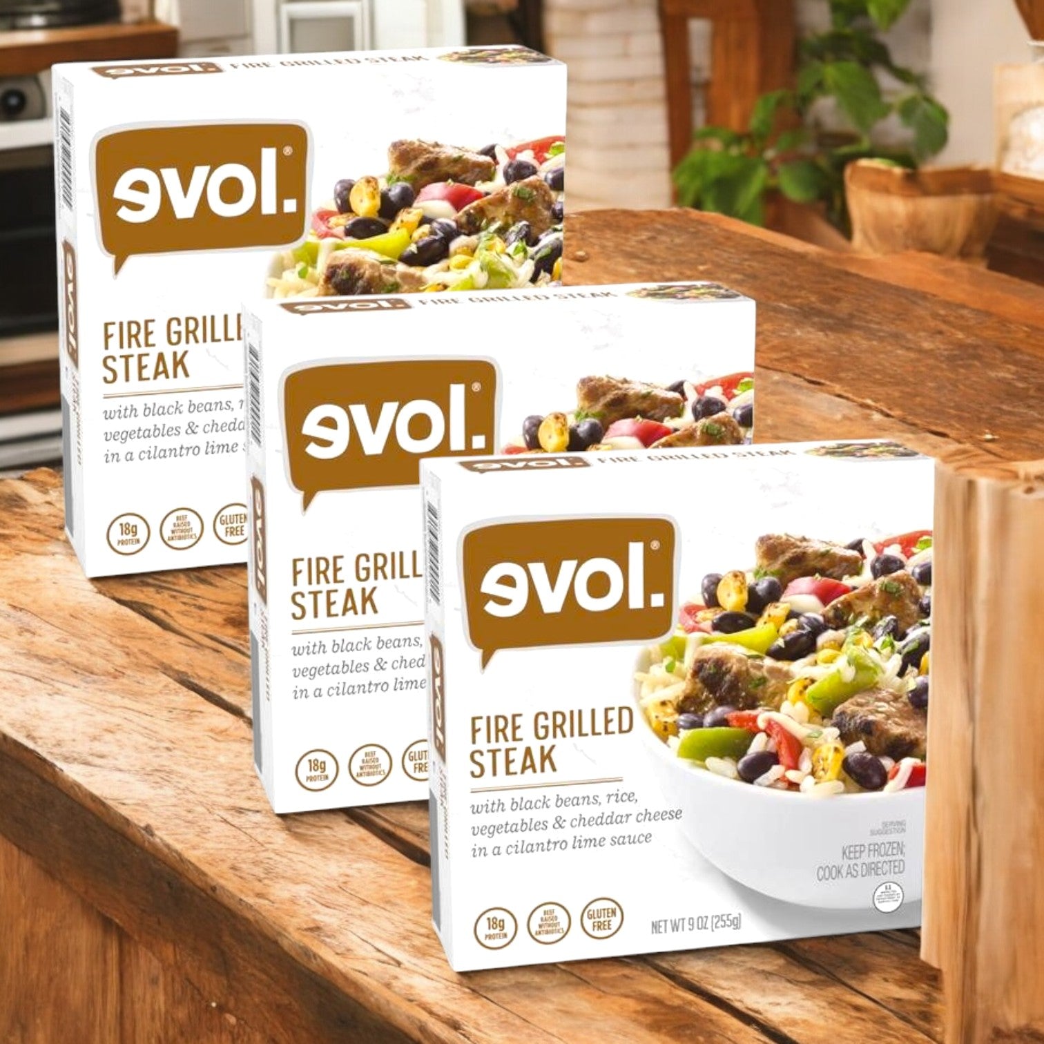 Three packs of Evol Gluten Free Fire Grilled Steak, 9 oz., are displayed on a wooden kitchen counter, showcasing a hearty meal with vegetables and rice pictured on the packaging. Enjoy this delicious option from Evol any night of the week!.