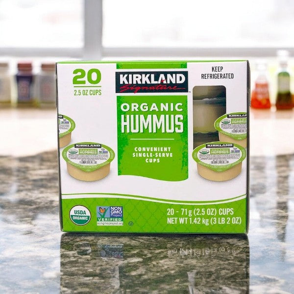 A box of Kirkland Signature Organic Hummus, featuring 20 single-serve cups made from premium organic chickpeas, sits gracefully on a marble countertop.