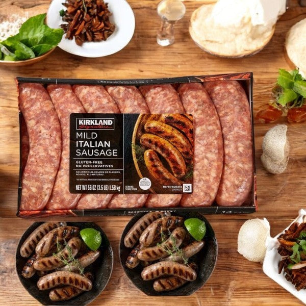 A Kirkland Signature Mild Italian Sausage-3 Count package with premium pork infused with Italian herbs is elegantly displayed alongside cooked sausages on plates, all set on a wooden table adorned with various ingredients.