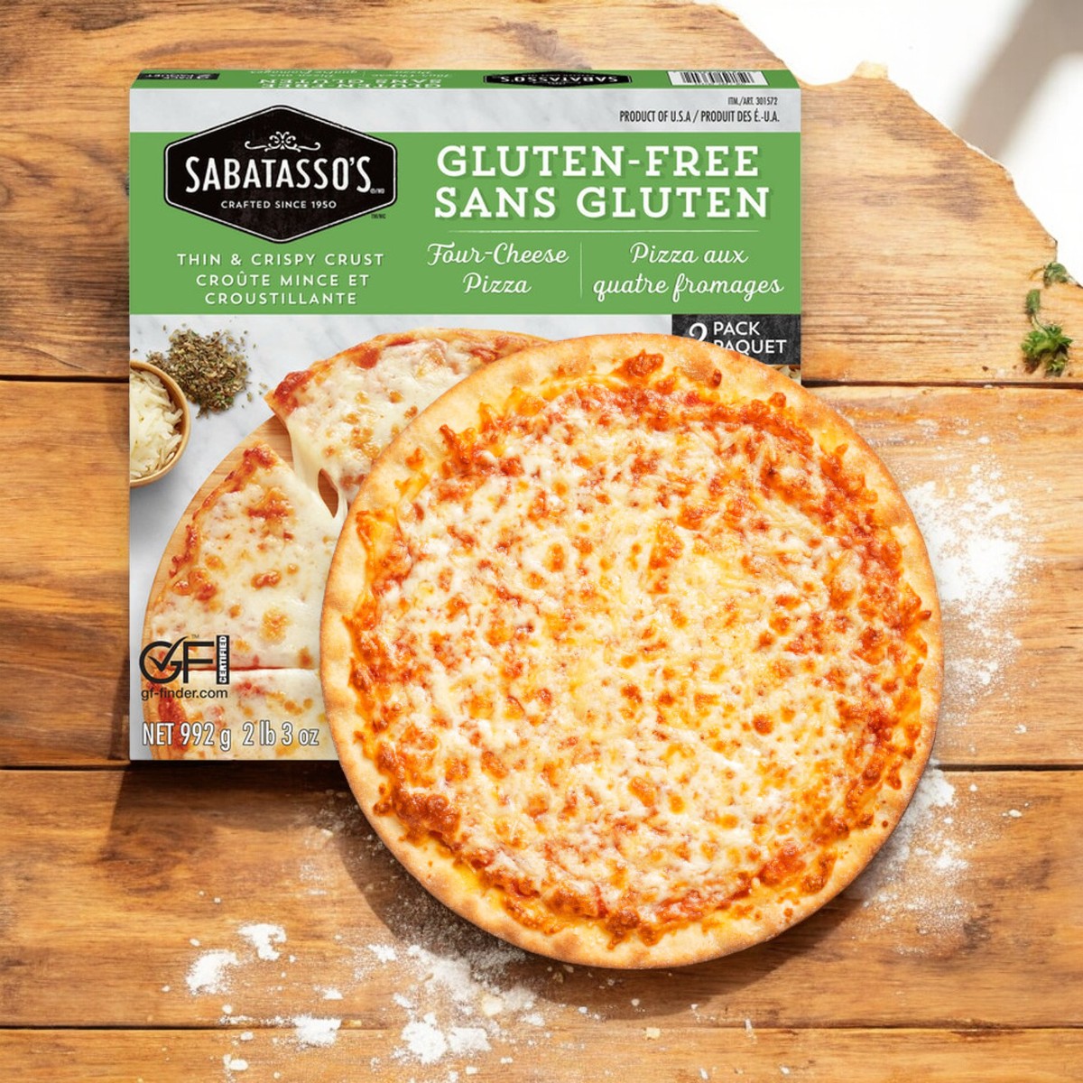 A 3-pack of Sabatasso's Gluten-Free Four Cheese Pizza sits beside a cooked pizza with a crispy crust, resting on a flour-dusted wooden surface.