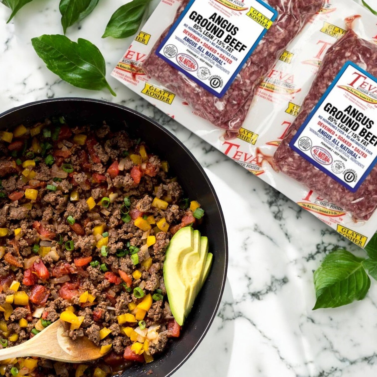 A skillet with cooked Angus ground beef, diced yellow peppers, and tomatoes is on a marble surface. Nearby are packs of Teva Kosher Foods Glatt Kosher 85% lean Angus ground beef and fresh basil leaves.