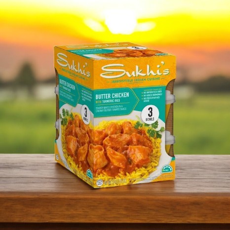 A box of Sukhi's Butter Chicken with Turmeric Rice, available in a 3 Pack-1 Case, offers an Indian-inspired meal. The background depicts a blurred sunset over fields, merging the warmth of tradition with modern convenience.