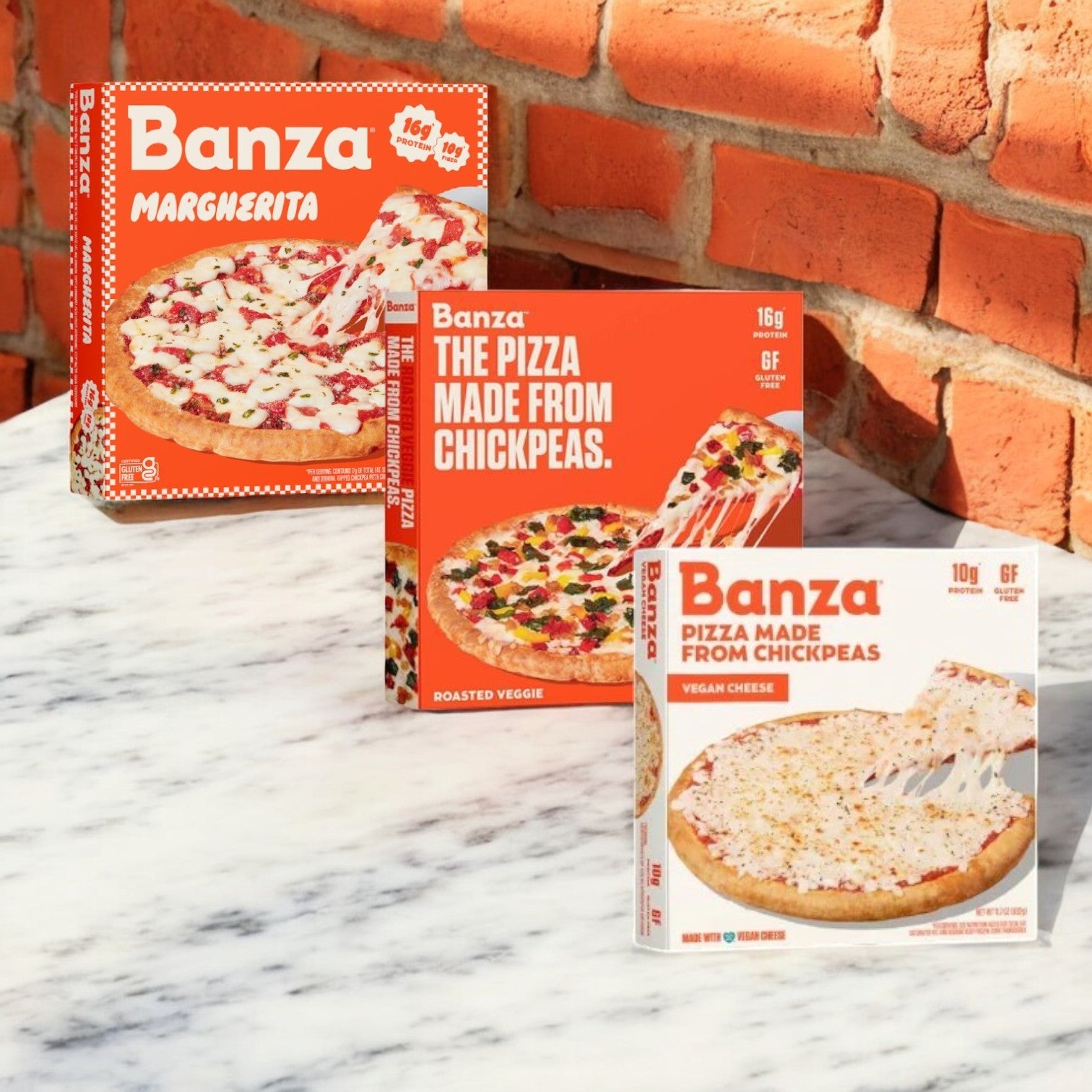 Three boxes of the Banza 3 Pack Combo! featuring flavors like Margherita, Roasted Veggie, and Vegan Cheese, are elegantly placed on a marble surface against a rustic brick wall.