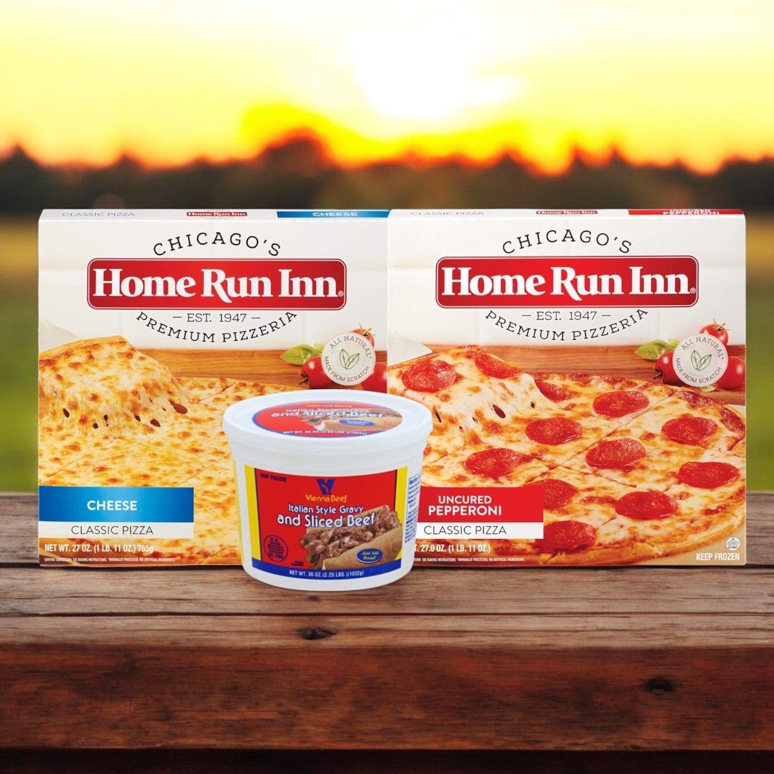 The Home Run Inn and Vienna Beef Chicago-Style Favorites Combo by Easy Lunches features two pizza boxes, cheese and uncured pepperoni, with a tub of sliced Italian beef on a wooden surface, set against a warm sunset.