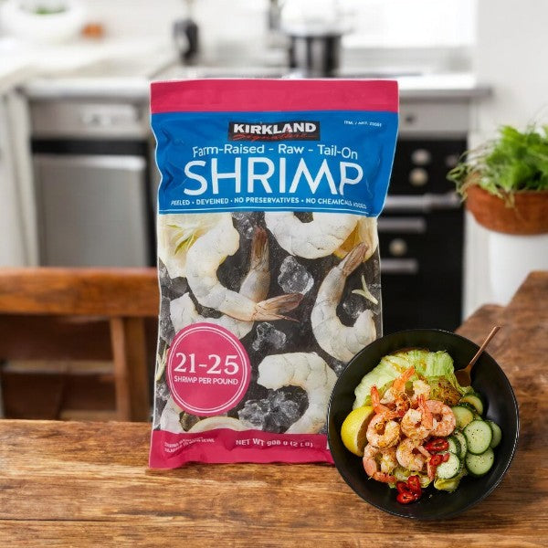 A bag of Kirkland Signature Farm-Raised Raw Shrimp, Tail-On, Peeled, Deveined (2 lbs) rests on a wooden table beside a colorful salad with shrimp, cucumbers, avocados, and tomatoes in a cozy home kitchen.