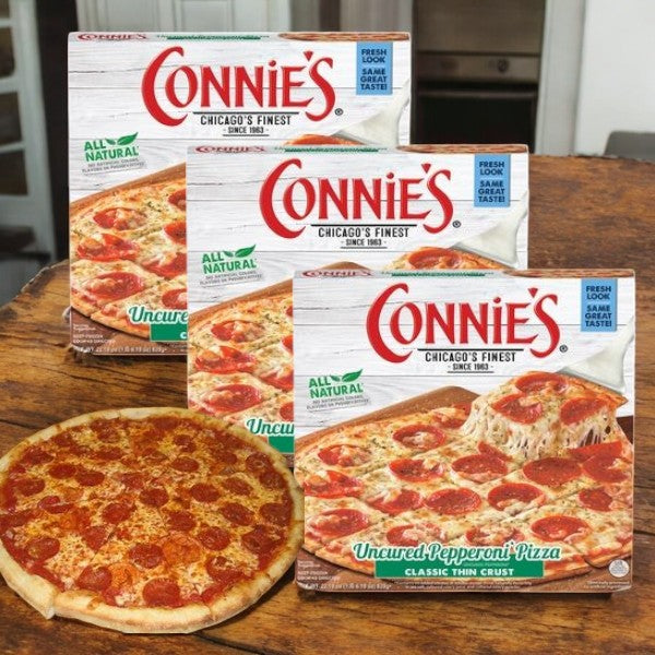 Three boxes of Connie's Pizza, Classic Thin Crust, Uncured Pepperoni (22.19 oz.) are displayed on a wooden table beside a baked pepperoni pizza, making for a quick and convenient meal from Connie's Pizza 3-Pack.