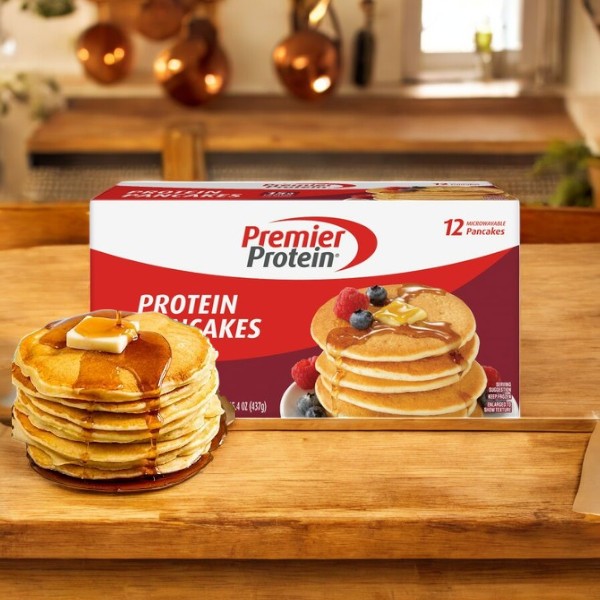 A box of Premier Protein Pancakes – 12 Count – 1 Pack is on the kitchen table next to a stack of Premier Protein pancakes topped with butter and syrup.