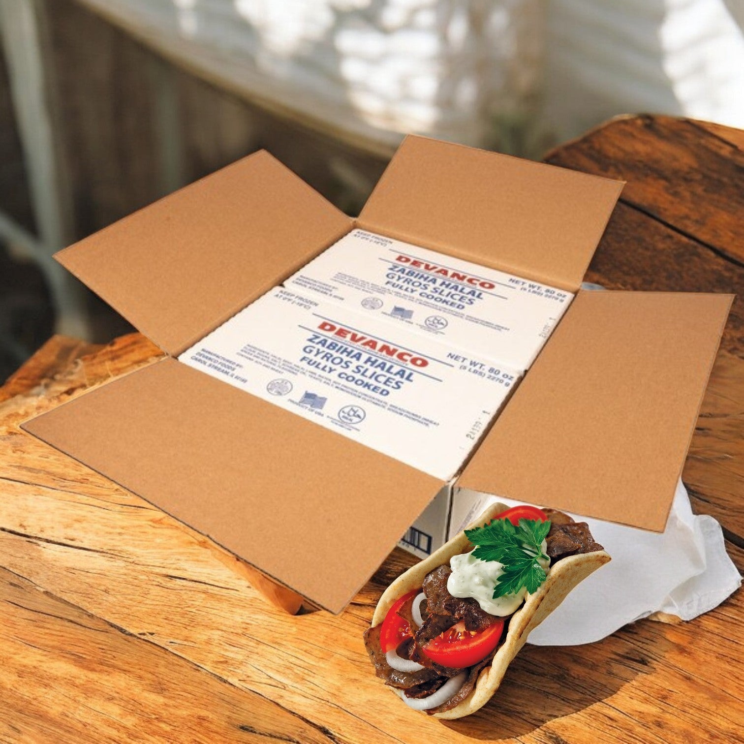 An open cardboard box with white packages rests on a wooden surface, while a pita sandwich bursting with Devanco's Halal Gyro Slices - 5 lb, seasoned with Mediterranean spices, tomatoes, onions, and parsley, lies enticingly beside it.